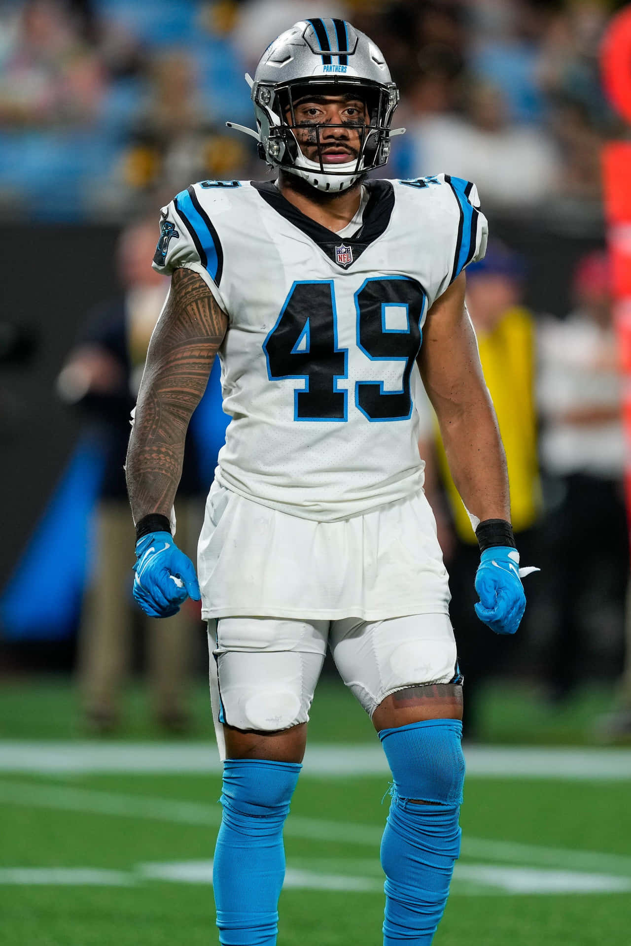  壁紙 — 一位專注的NFL線衛身穿白色和藍色制服，站在綠色球場上，身後體育場燈光閃爍。