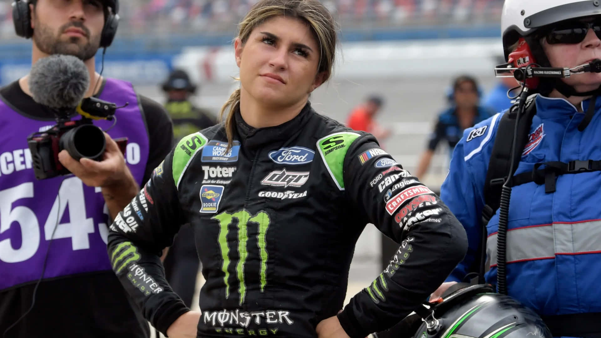  壁紙 — 一位專注的女性NASCAR車手穿著黑色和綠色的Monster Energy賽車服，雙手叉腰站在賽道上。