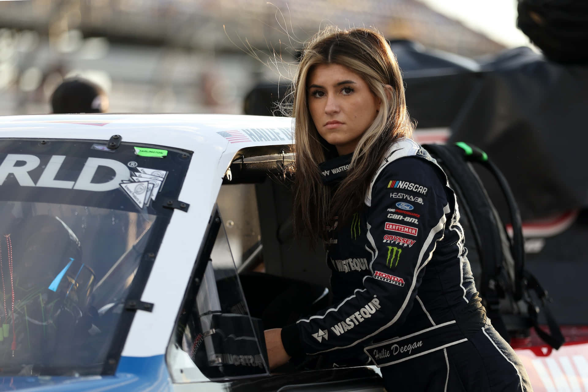  壁紙 — 一位專注的女性NASCAR車手身穿帶有贊助商標誌的品牌賽車服，從她的賽車中探出身子。