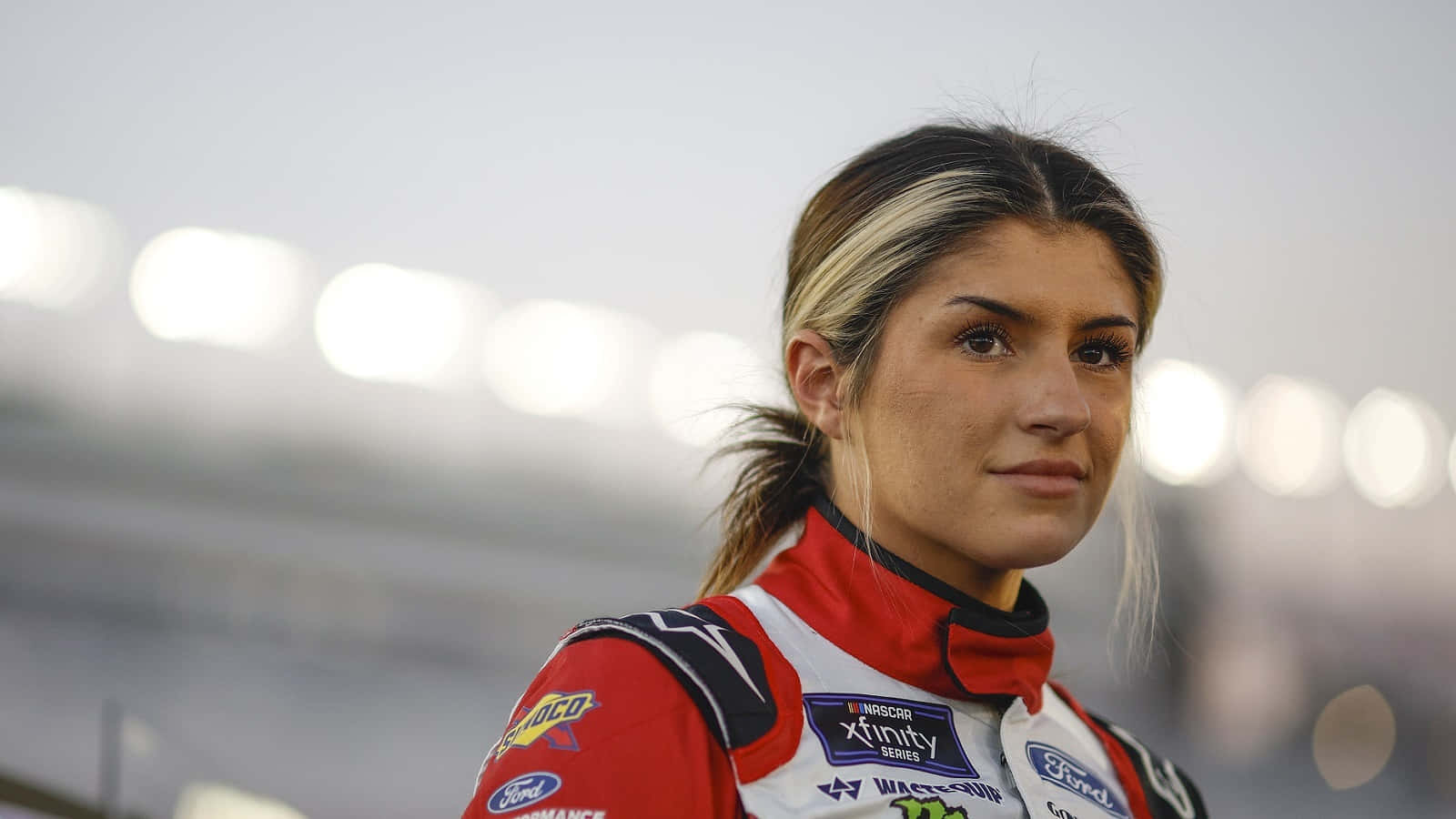  壁紙 — 一位專注的女性NASCAR車手，身穿紅白賽車服，在模糊的體育場背景下堅定地向前凝視。