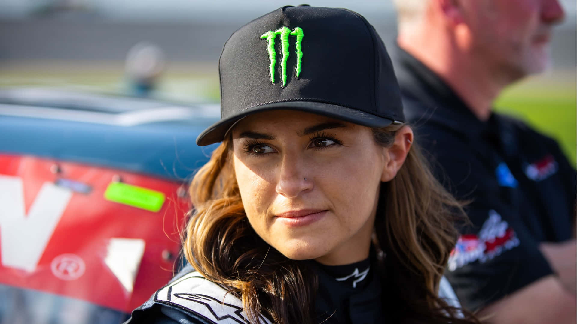  壁紙 — 一位專注的女性賽車手，戴著黑色Monster Energy帽子和賽車服，站在賽道上的車輛旁。