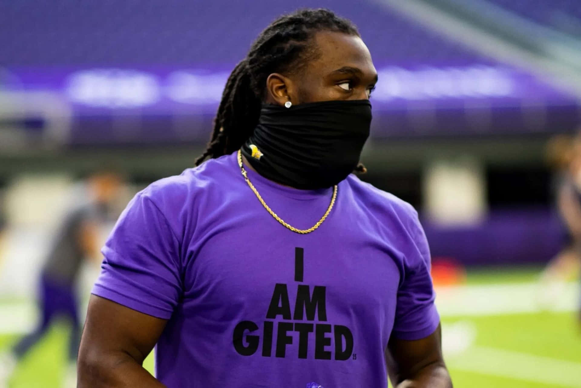  壁紙 — 一位專注的運動員穿著紫色 'I AM GIFTED' 襯衫和黑色面罩，站在體育場燈光下的綠色足球場上。