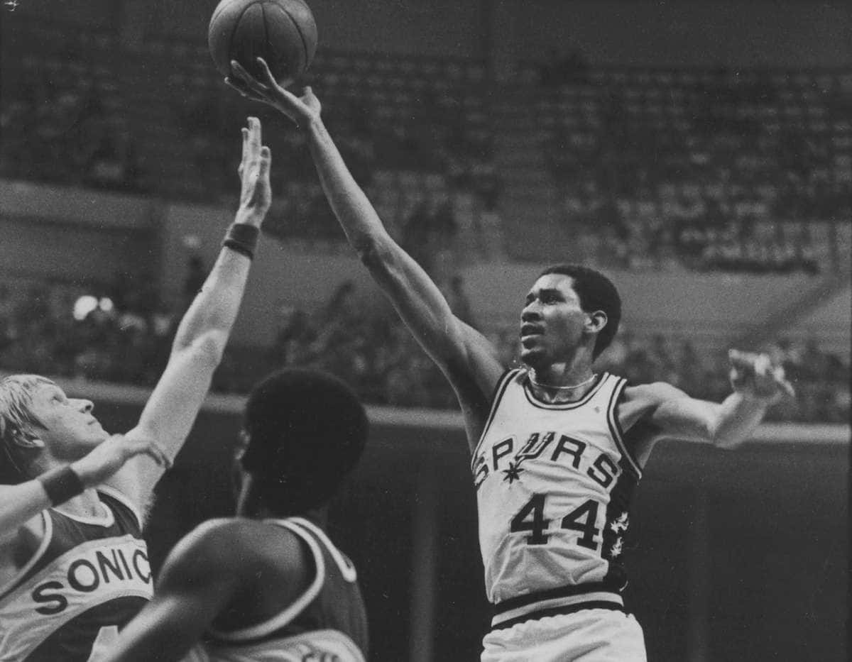 身穿馬刺隊球衣的籃球運動員在比賽中越過防守者投籃。 — NBA 壁紙