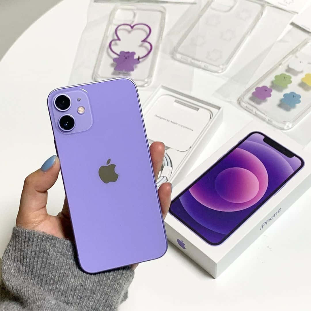 手拿著紫色 iPhone 12 mini，附近有包裝和雲朵主題保護殼。 — iPhone 12 壁紙