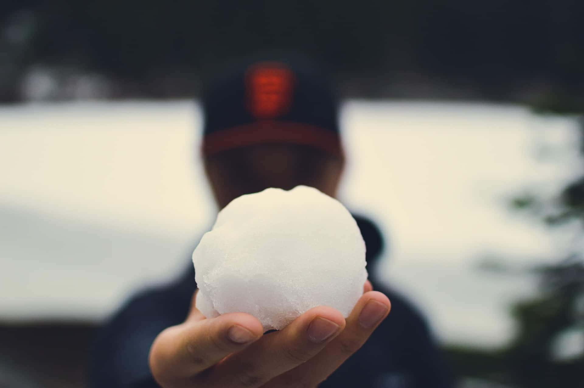  壁紙 — 一隻手拿著一個完美的雪球，焦點清晰，而背景中一個戴著帽子的人模糊地站在雪景中。