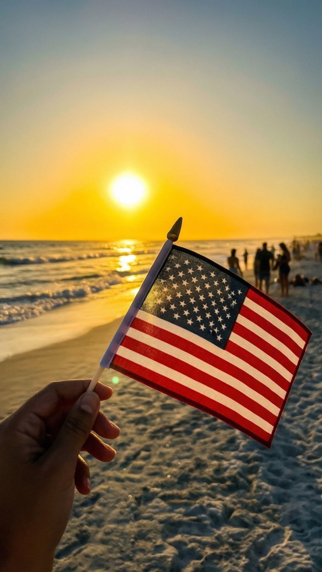 Flag at Sunset 壁紙 — 一隻手拿著一面小型美國國旗，背景是金色日落下的沙灘，遠處有人群。