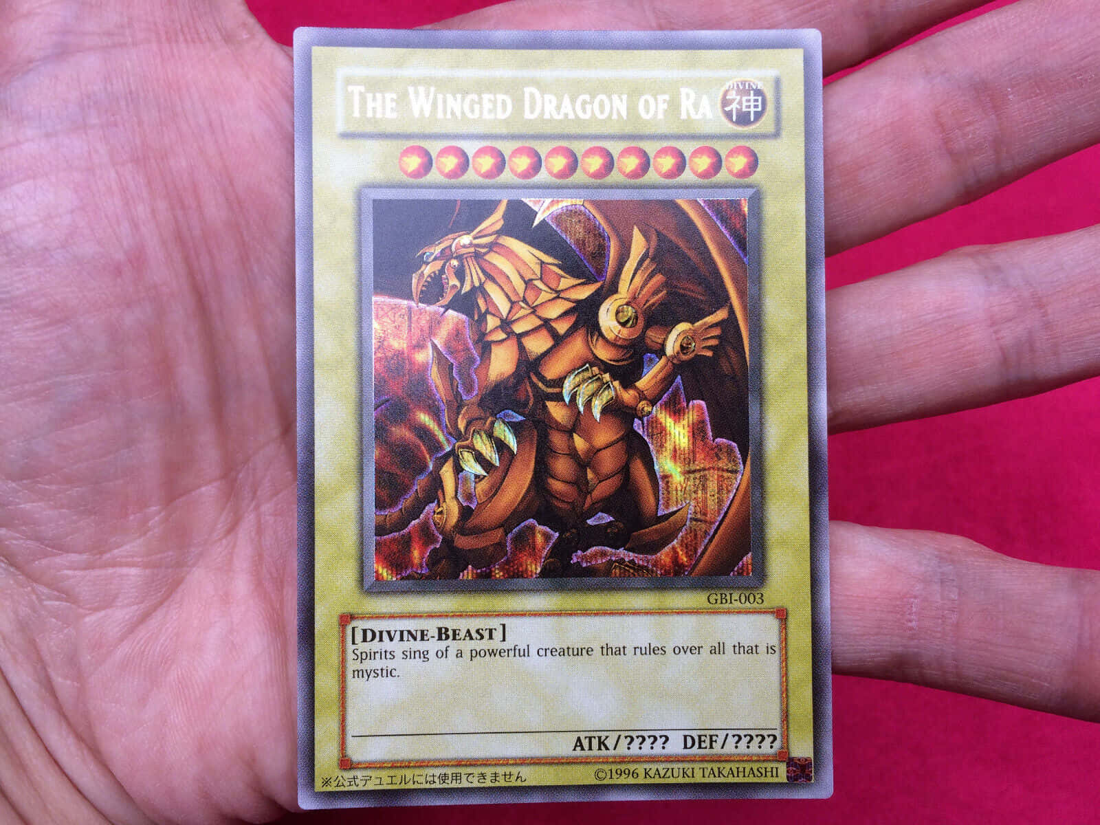 The Winged Dragon Of Ra 桌布 壁紙 — 一隻手拿著一張印有「拉的翼神龍」的遊戲王交換卡，背景是鮮紅色。