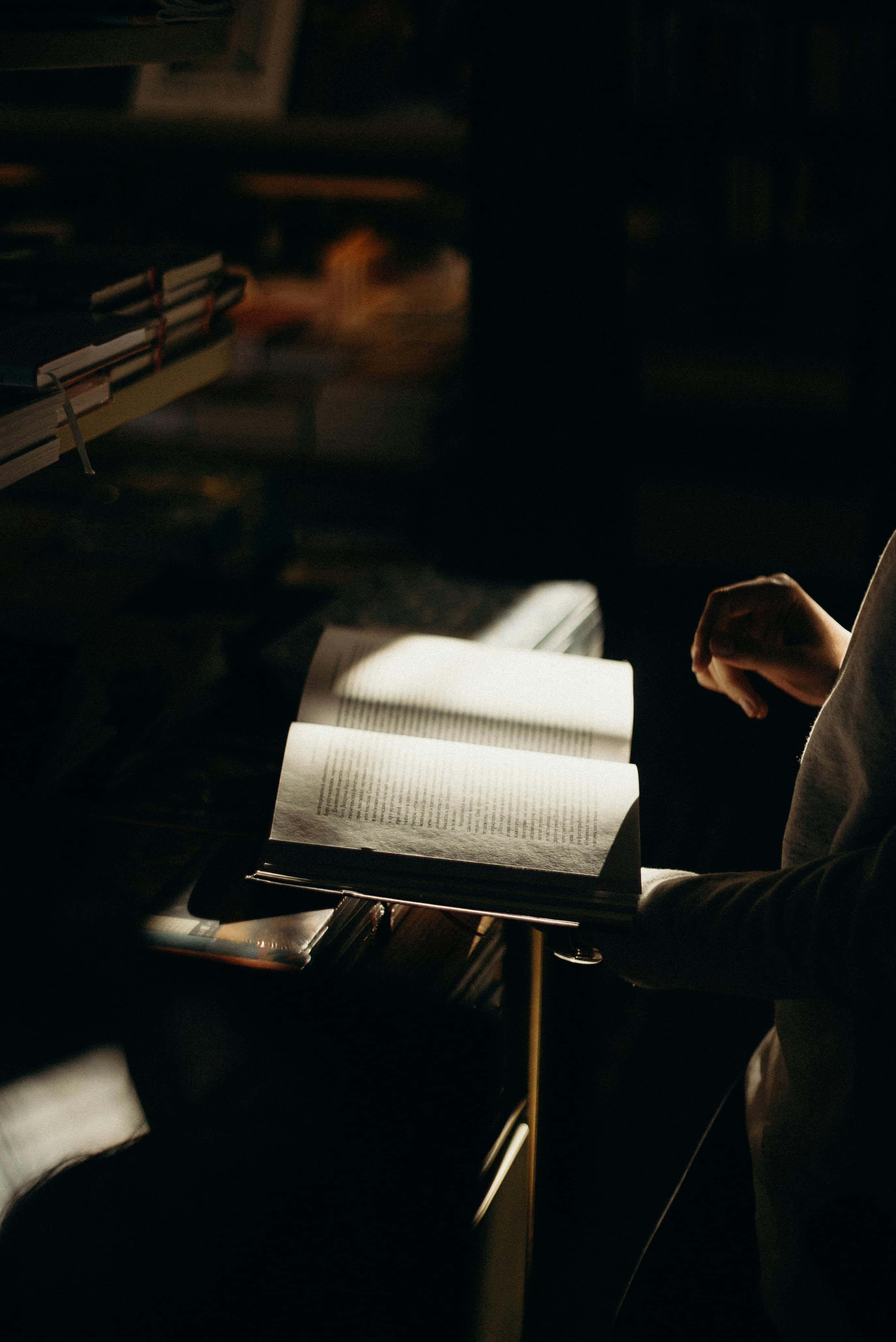 Dark Book Aesthetic 桌布 壁紙 — 一隻手拿著一本打開的書，沐浴在溫暖的光線中，背景是一個充滿書架的黑暗房間。