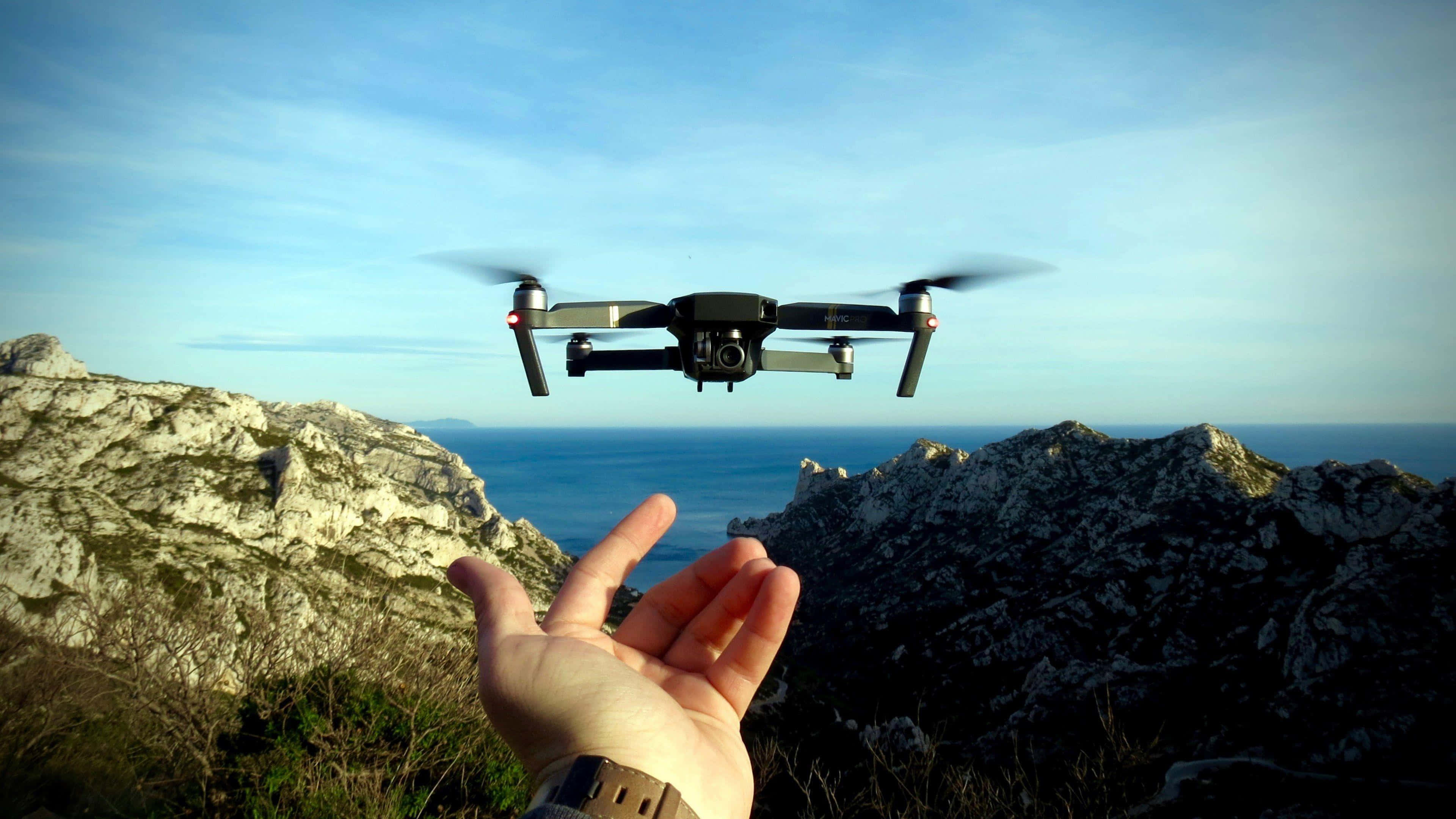 Drone Camera 桌布 壁紙 — 一隻手在晴朗藍天下，於崎嶇懸崖和海洋上方釋放一架懸停的無人機。