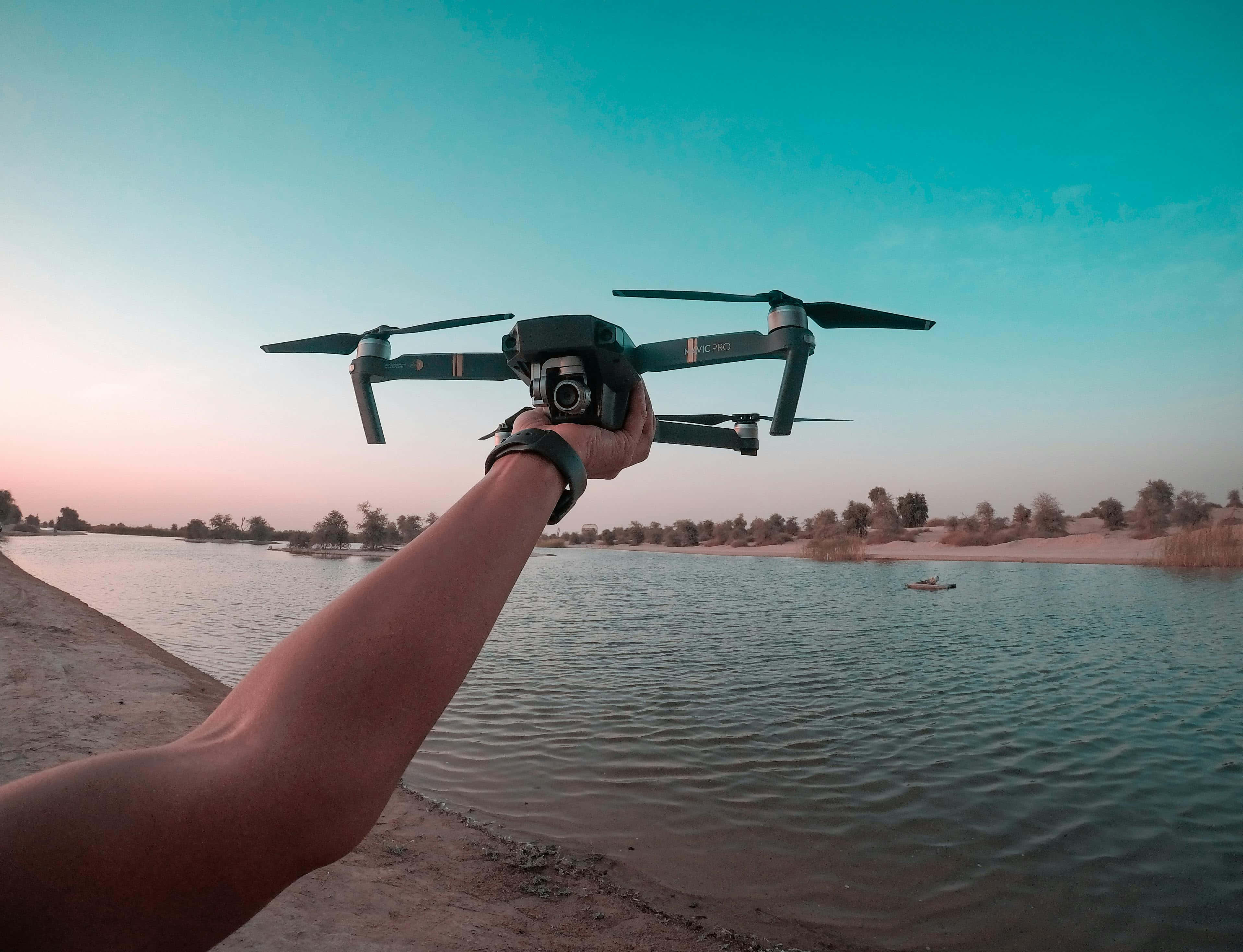 Drone Camera 桌布 壁紙 — 一隻手在日落時分，粉紅與青綠色的天空下，將無人機舉在平靜的湖面上。