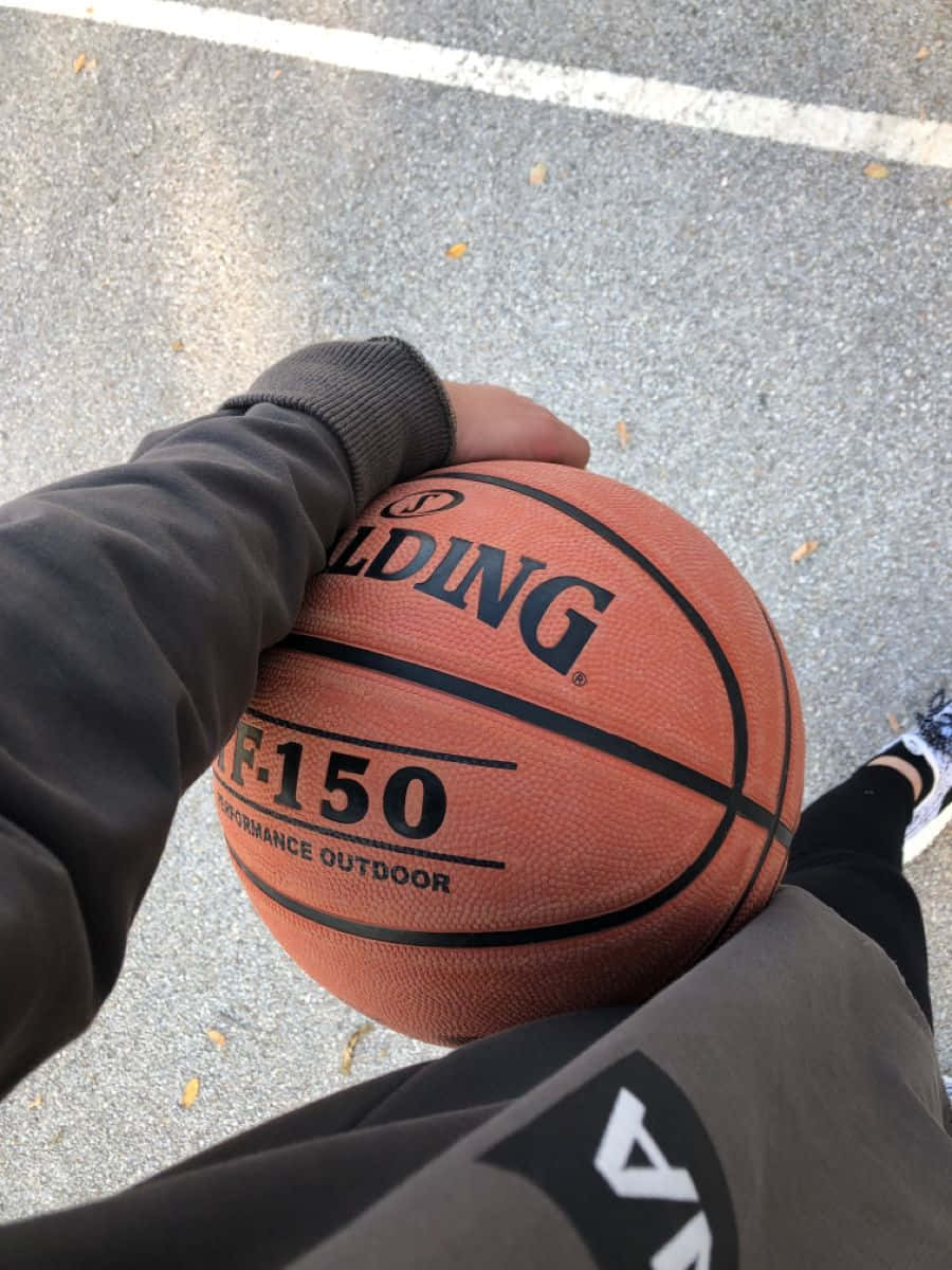  壁紙 — 一隻手抓住一個橙色 Spalding 籃球，上面標有 F-150 Performance Outdoor，在灰色瀝青球場上，有白色線條。