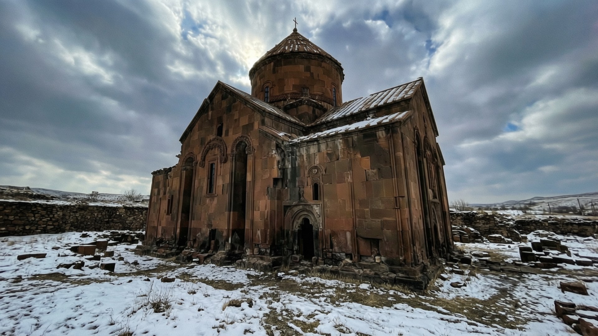 Ani Ruins Winter 壁紙 — 一座被雪覆蓋的古老石教堂，圓頂屋頂，在戲劇性的多雲天空下莊嚴地矗立。