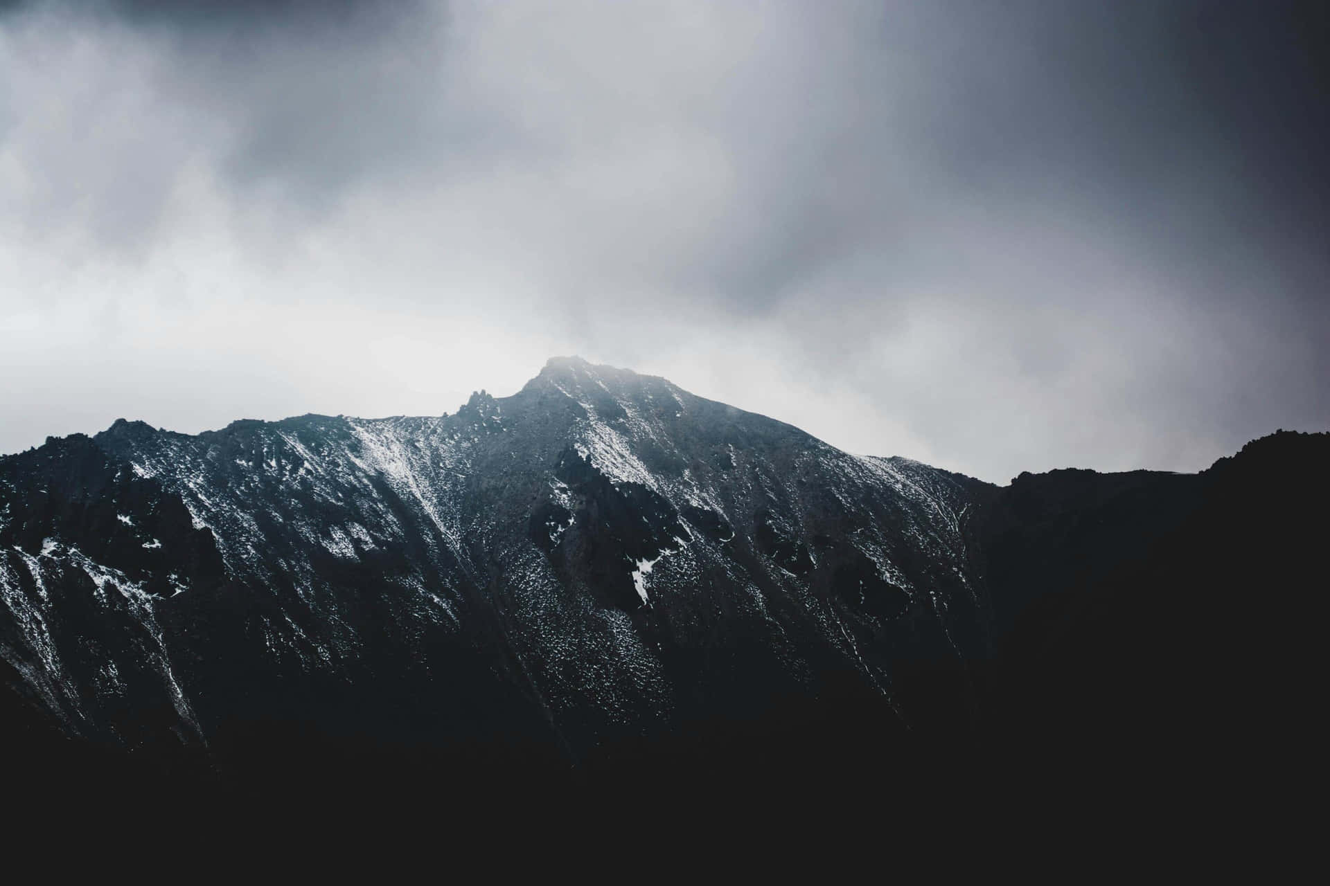  壁紙 — 一座陡峭的山峰刺穿厚重的灰色雲層，白雪依附在其崎嶇的山脊上，光線昏暗。