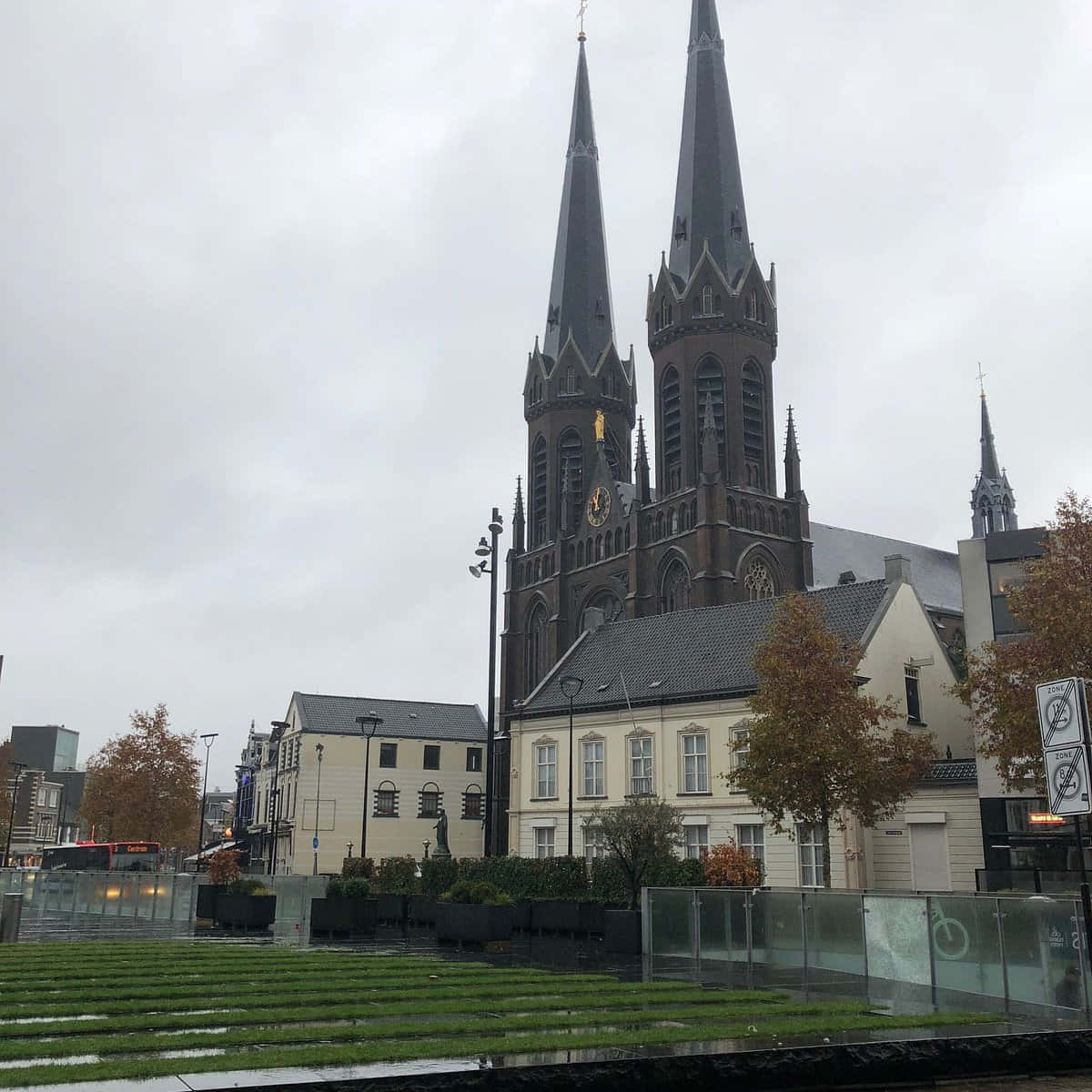 Tilburg 桌布 壁紙 — 一座宏偉的哥德式大教堂，擁有雙尖塔，聳立在濕潤的城市廣場上，廣場兩旁種滿了秋樹和現代玻璃欄杆，天空多雲。