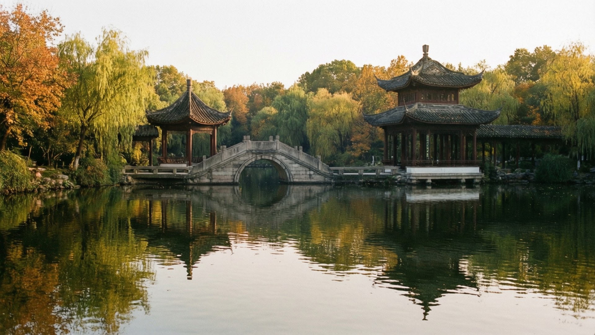 Luhu Lake Autumn 壁紙 — 一座石拱橋連接兩座傳統中式亭閣，倒映在平靜的水面上，周圍是秋樹。