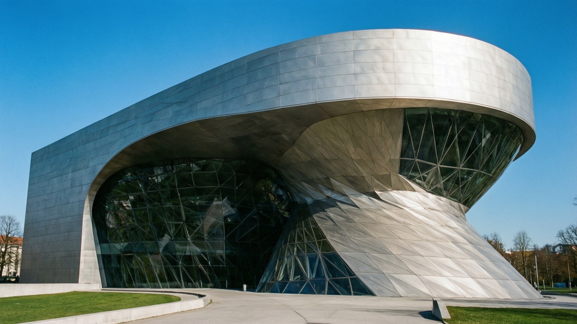 BMW Museum Modernity 壁紙 — 一座未來風格的銀色玻璃建築在晴朗的藍天下戲劇性地彎曲，下方是修剪整齊的綠色草坪。