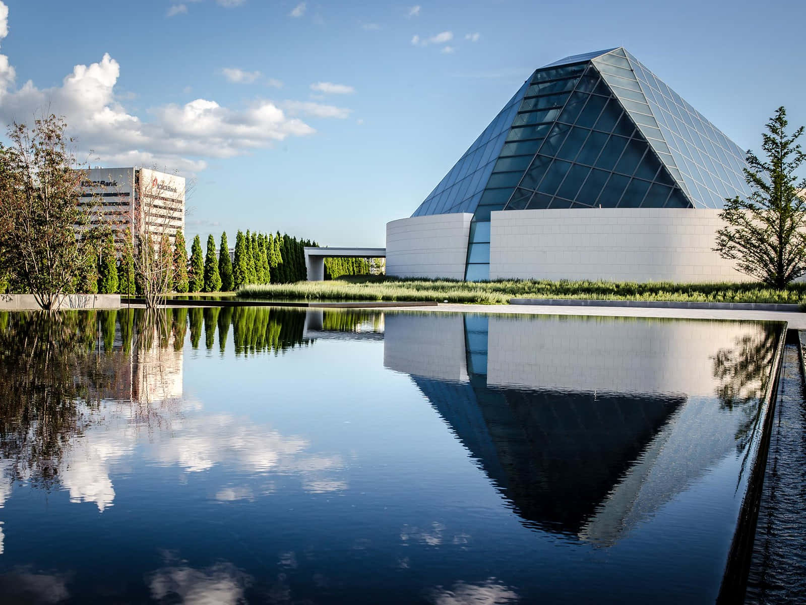 Aga Khan Museum 桌布 壁紙 — 一座現代玻璃金字塔建築在晴朗藍天和零星白雲下，完美倒映在平靜的池塘中。