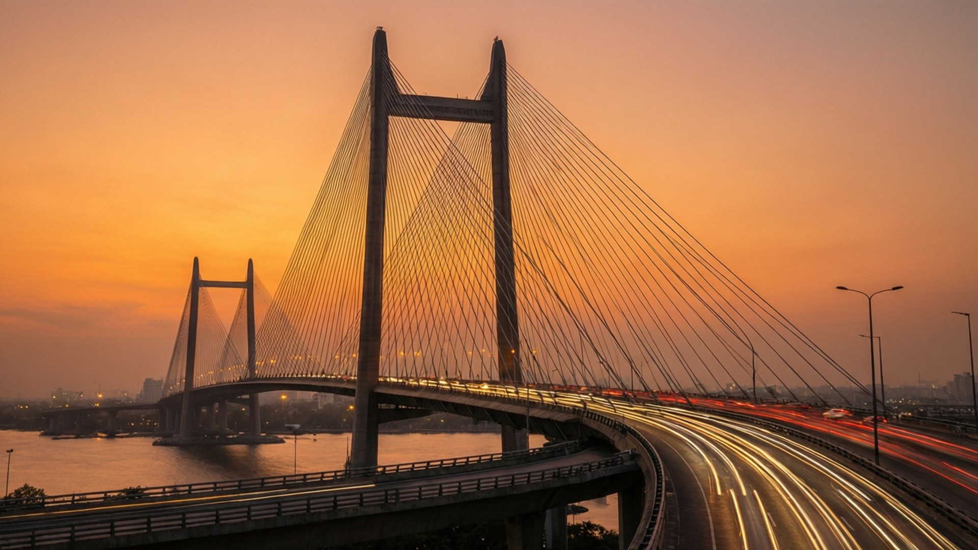 Sea Link Sunset 壁紙 — 一座斜張橋在日落時跨越河流，移動車輛的光跡營造出動感的城市景象。