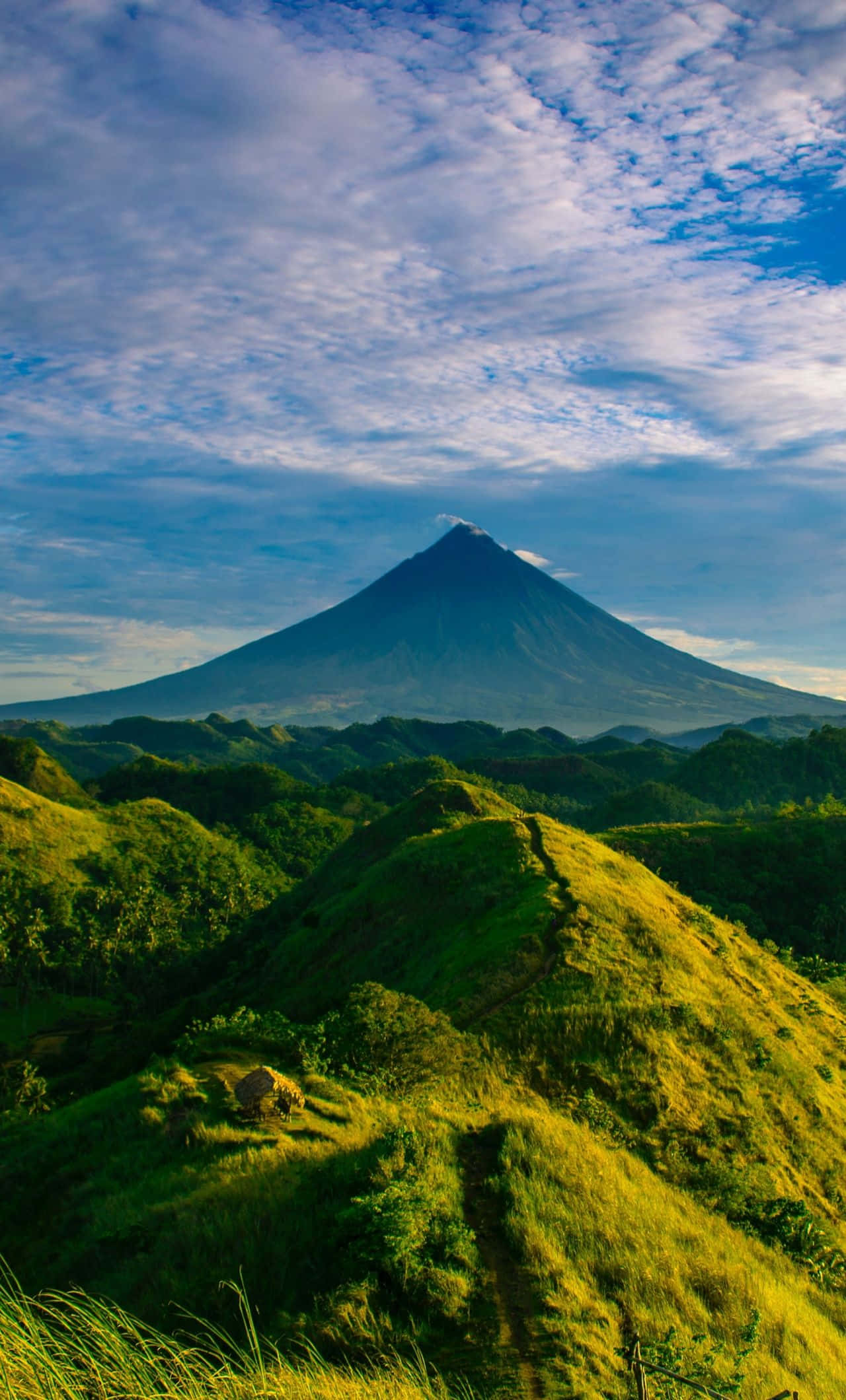 Hill 桌布 壁紙 — 一座雄偉的圓錐形火山在蔚藍天空和縷縷白雲下，聳立於起伏的綠色山丘之上。