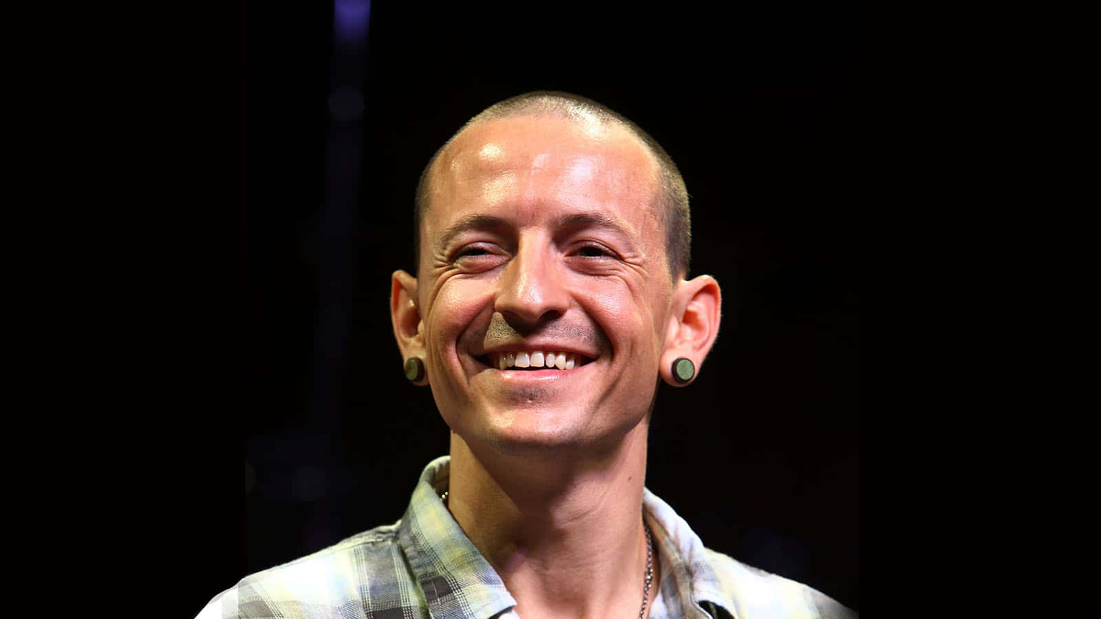  壁紙 — 一個光頭並戴著綠色耳擴的男子，在深色背景下洋溢著喜悅。