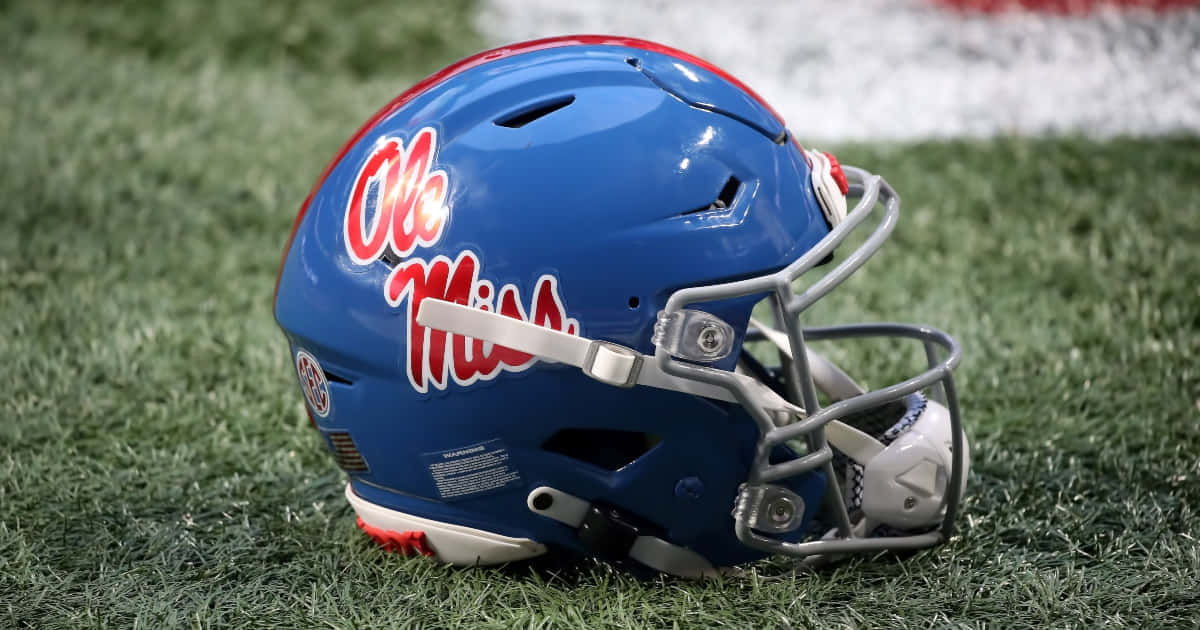  壁紙 — 一個帶有紅色字樣的藍色 Ole Miss 美式足球頭盔放在白色邊界線附近的綠色草地上。