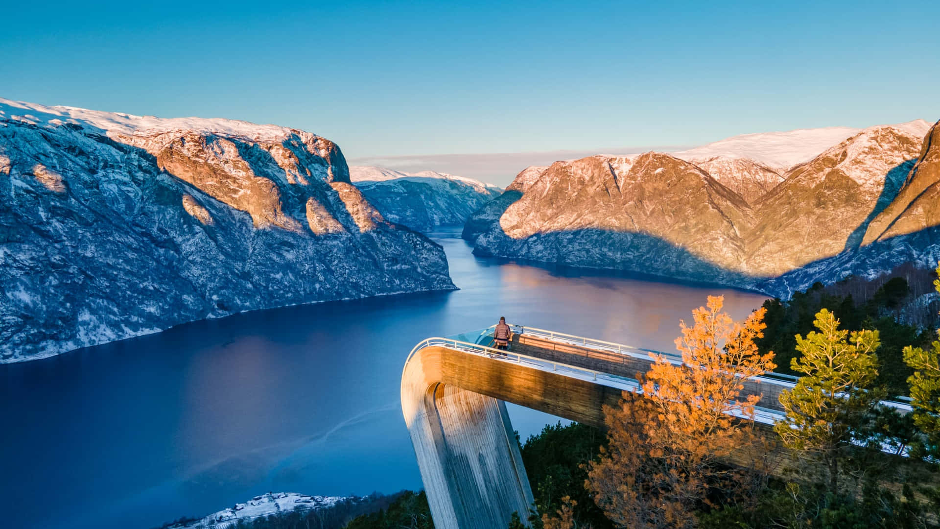  壁紙 — 一個孤獨的身影坐在挪威 Trollstigen 的標誌性橋樑上，俯瞰著被積雪覆蓋的山脈環繞的寧靜峽灣，在清澈的藍天下。
