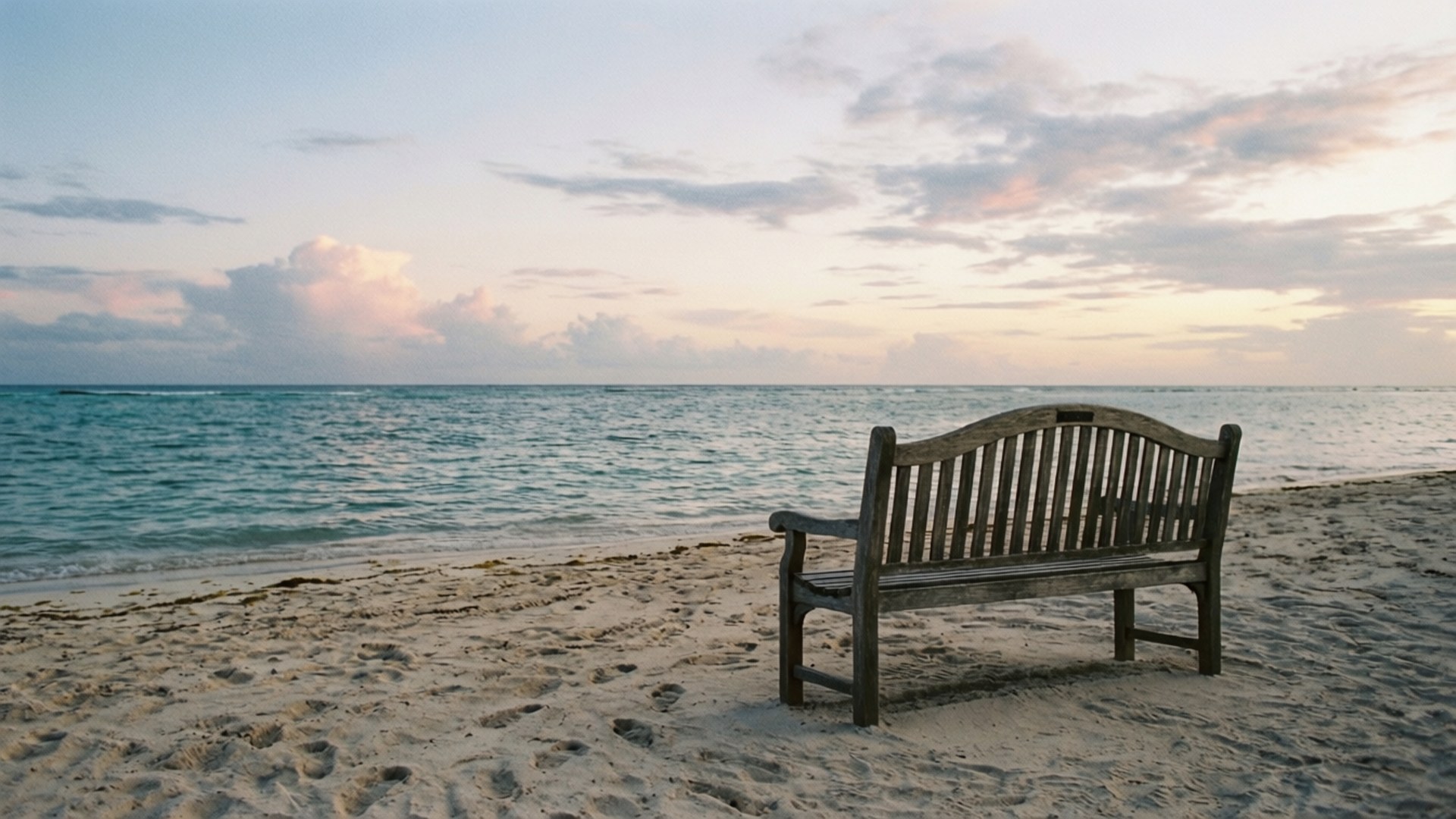 Morning Bench by the Sea 壁紙 — 一張風化的木長椅獨自坐在沙灘上，面向平靜的碧綠海水，上方是柔和粉彩的日落天空。