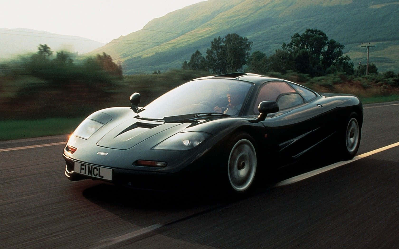 Mclaren F1 桌布 壁紙 — 一架流線型黑色McLaren F1在蜿蜒的山路上飛馳，背景是模糊的綠色山丘。