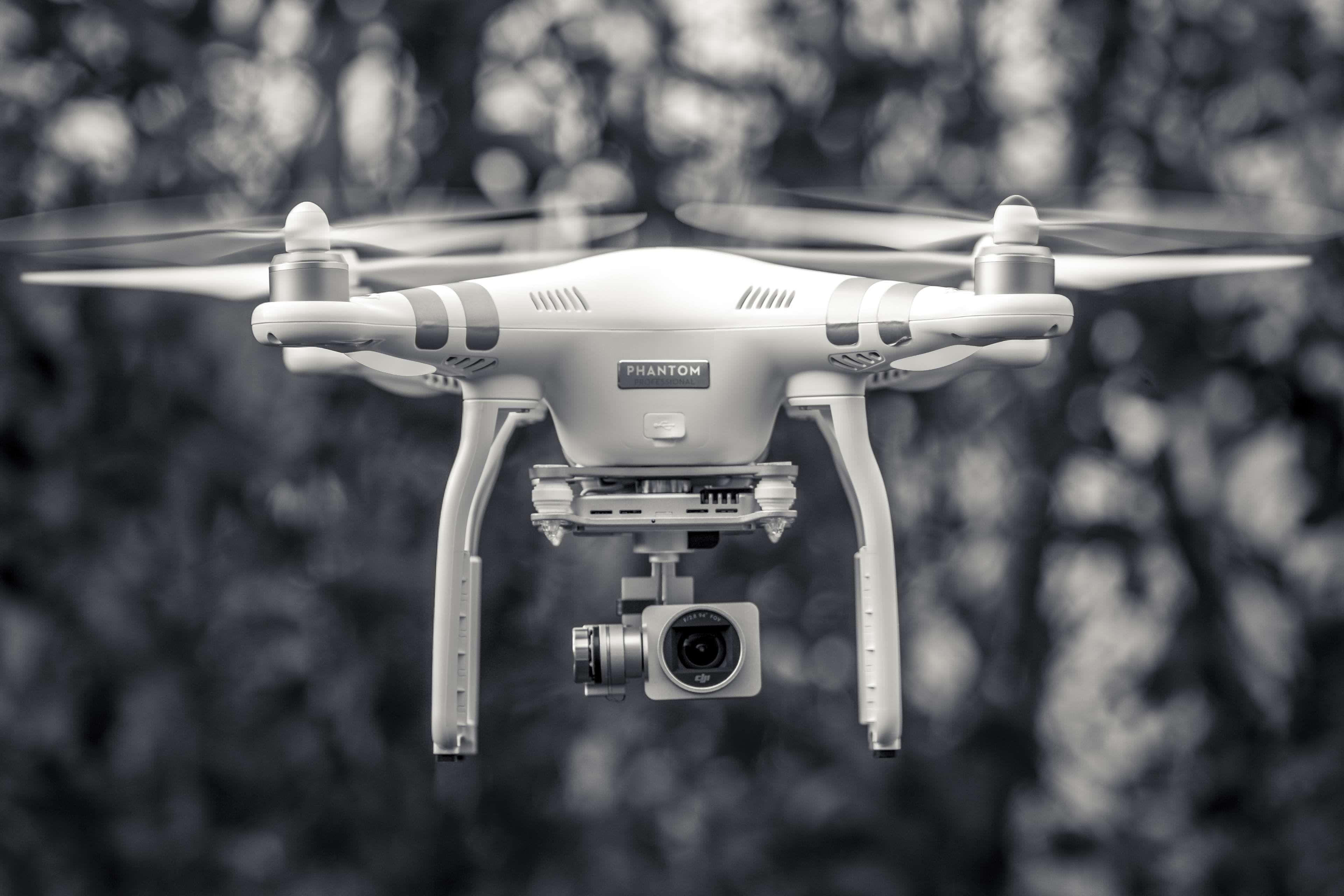 Drone Camera 桌布 壁紙 — 一架單色無人機在空中盤旋，旋翼轉動，背景是模糊的森林。