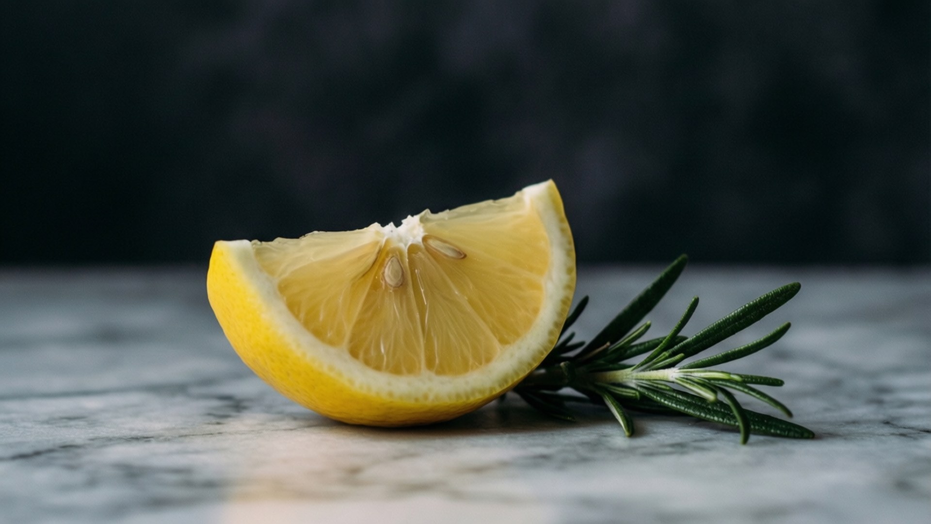 Elegant Lemon and Rosemary 壁紙 — 一塊鮮黃色的檸檬角和一枝迷迭香放在灰色大理石表面上，背景是深色模糊的。