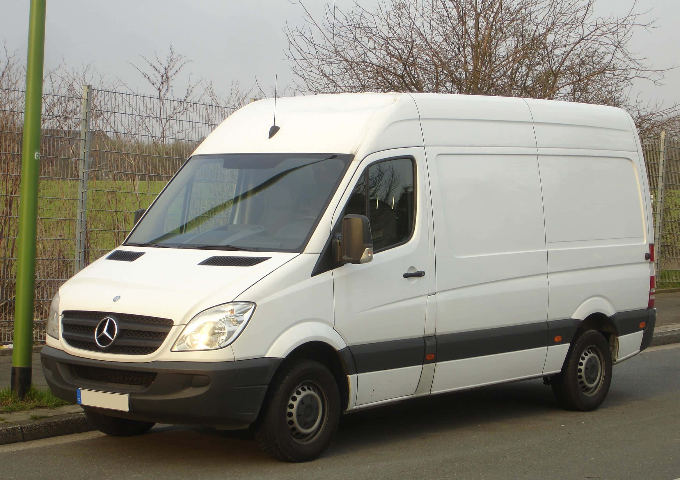 Mercedes Benz Sprinter 桌布 壁紙 — 一輛白色 Mercedes-Benz Sprinter 廂型車停放在街道上，旁邊有圍欄，背景有光禿禿的樹木。