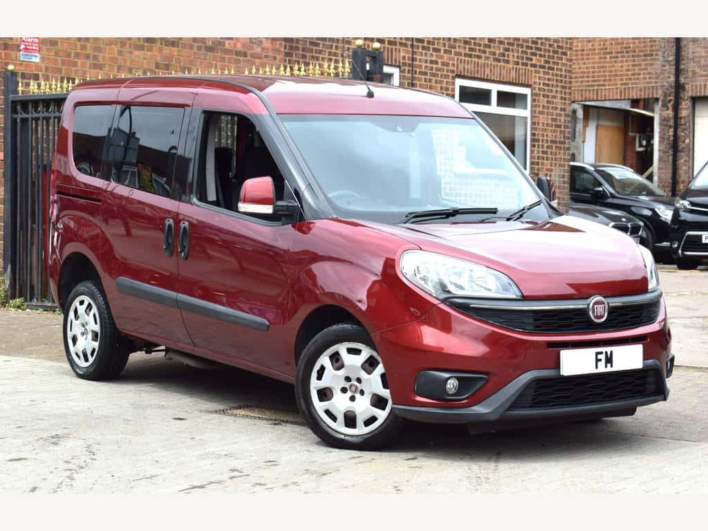  壁紙 — 一輛亮紅色的Fiat Fiorino廂型車停放在磚砌建築物前的鋪砌場地上，建築物設有黑色欄杆。