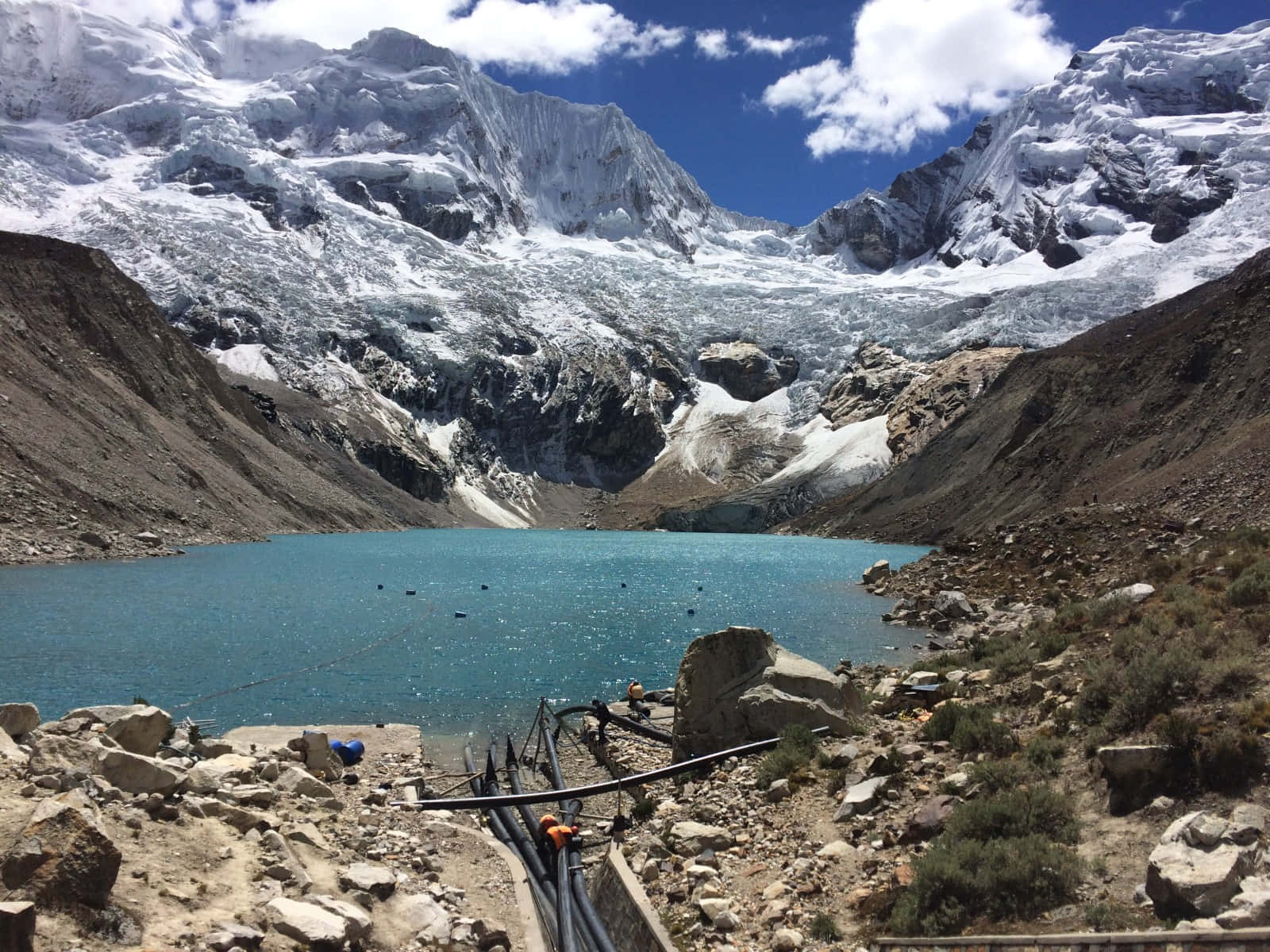  壁紙 — 一個令人驚嘆的綠松石冰川湖，坐落在崎嶇的雪山之中，上方是明亮的藍天和蓬鬆的白雲。