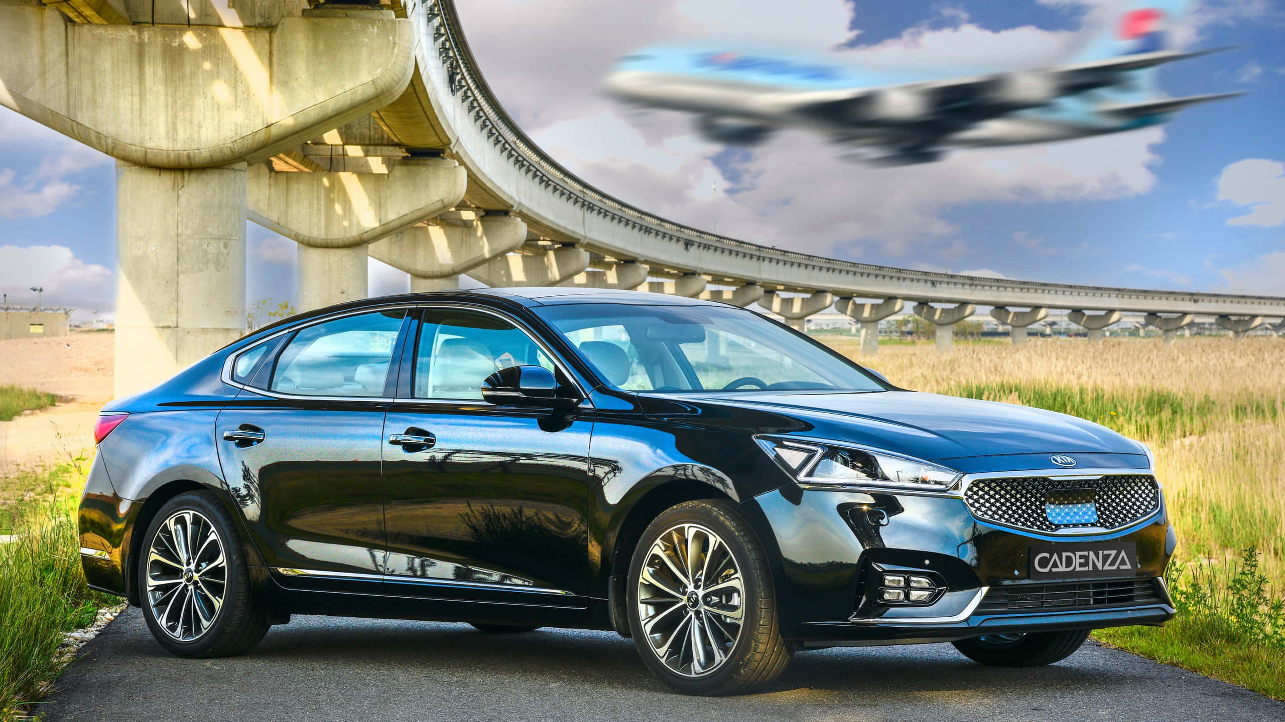 Kia Cadenza 桌布 壁紙 — 一輛時尚的黑色Kia Cadenza轎車停泊在彎曲的高速公路天橋下，一架模糊的飛機在上空飛過。