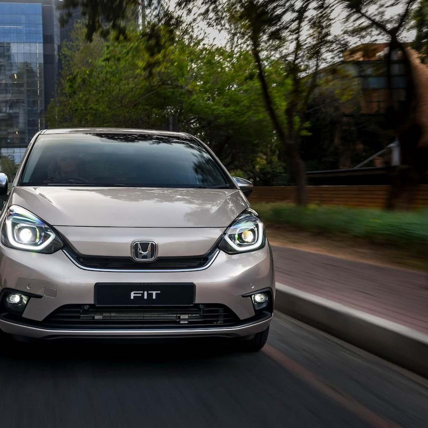 Honda Fit 桌布 壁紙 — 一輛銀色本田Fit在模糊的綠樹和現代建築背景下，於城市街道上向前行駛。