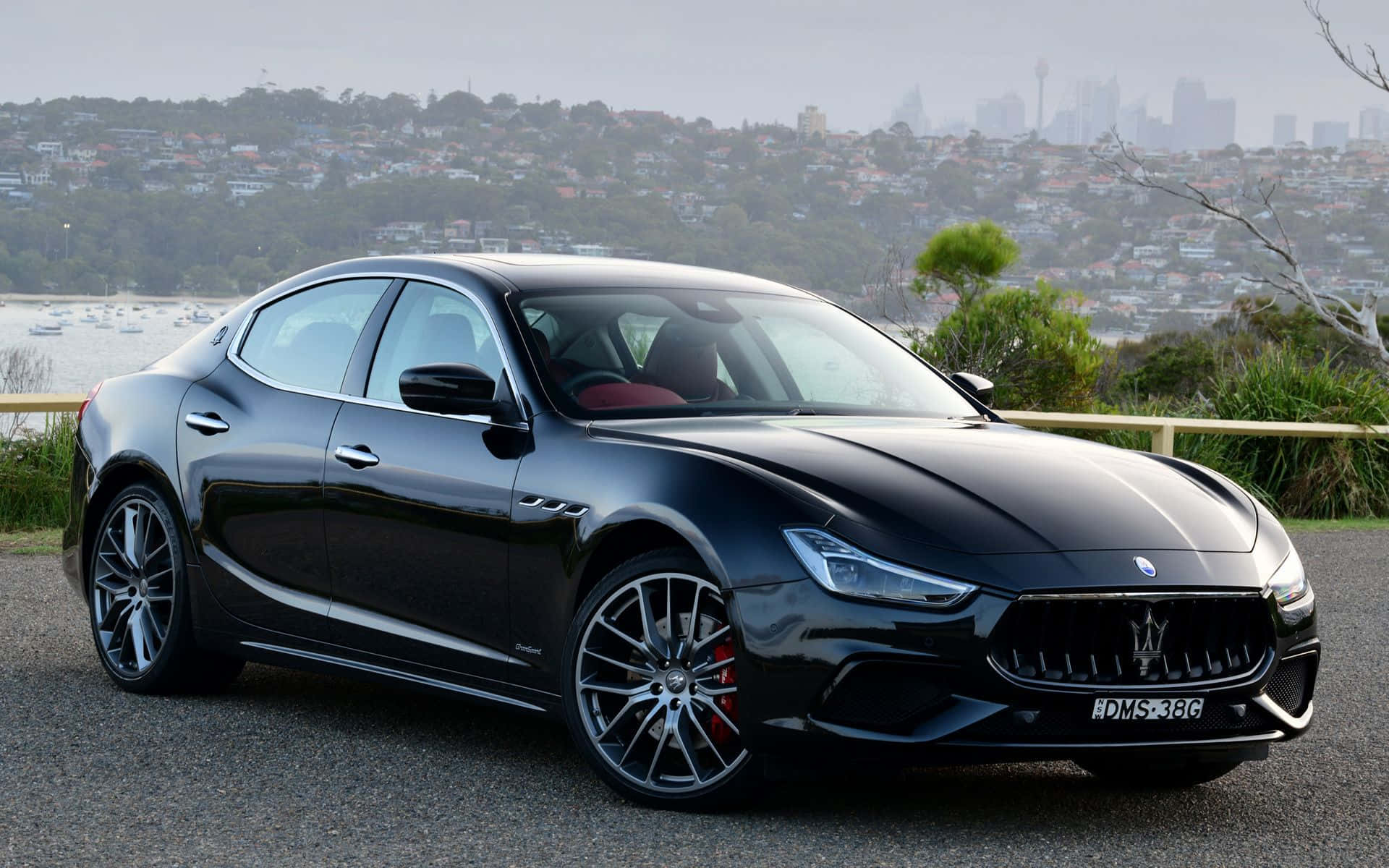 Maserati Ghibli 桌布 壁紙 — 一輛時尚的黑色瑪莎拉蒂Ghibli停泊在碎石觀景點上，背景是霧氣瀰漫的城市天際線和港口。