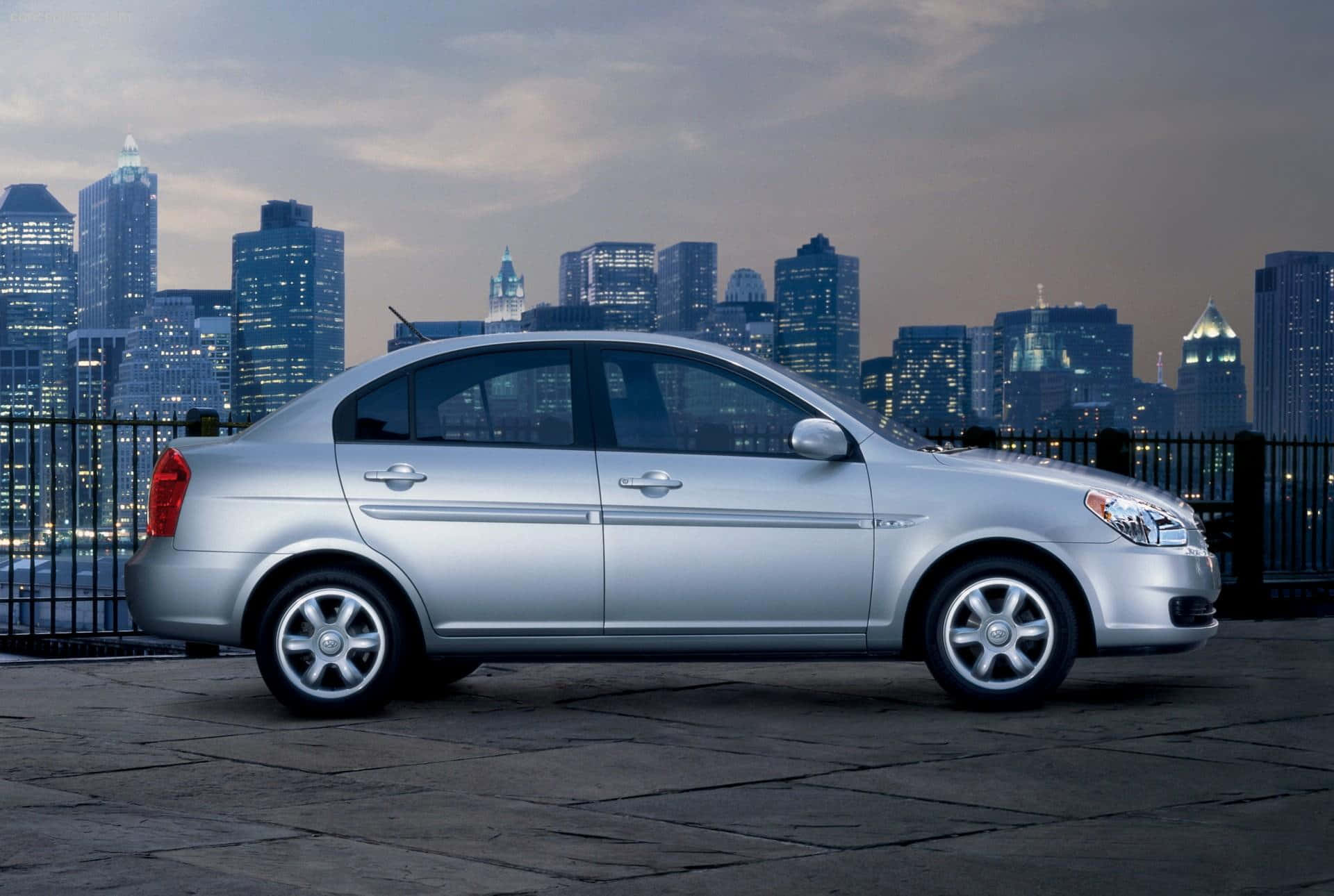 Hyundai Accent 桌布 壁紙 — 一輛銀色轎車停放在石屋頂上，俯瞰黃昏時分閃爍的城市天際線。