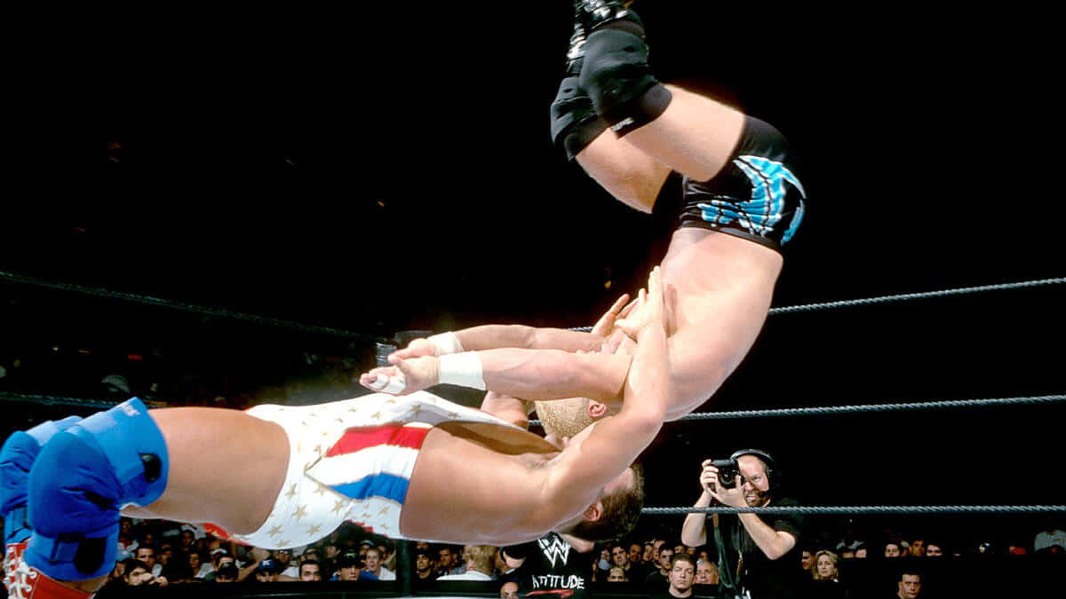 Crash Holly 桌布 壁紙 — 一名穿著藍色短褲的摔角手在燈光明亮的擂台上，以高飛動作跳過另一名穿著星條旗服裝的摔角手。