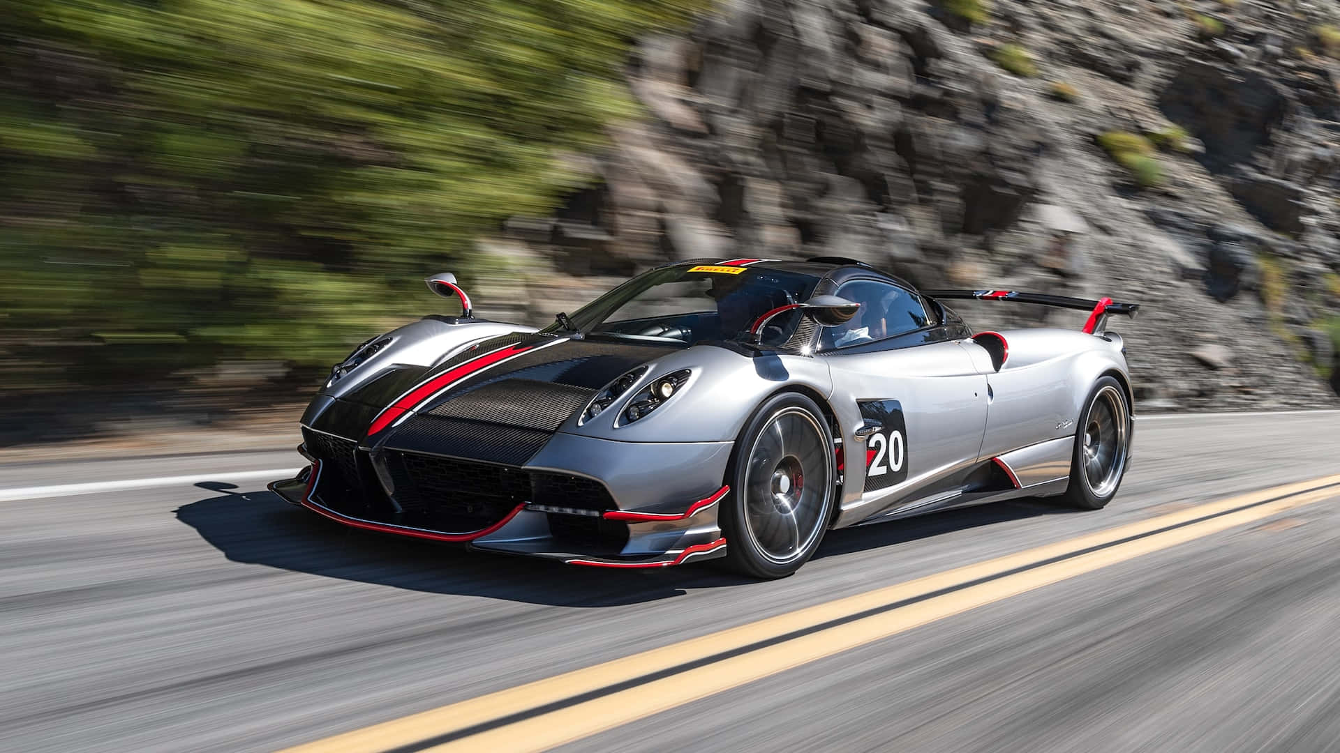 Pagani Huayra Roadster Bc 桌布 壁紙 — 一輛銀黑配色的帕加尼 Huayra BC 賽車，帶有紅色點綴，在蜿蜒的山路上疾馳，背景是模糊的綠樹和岩石峭壁。