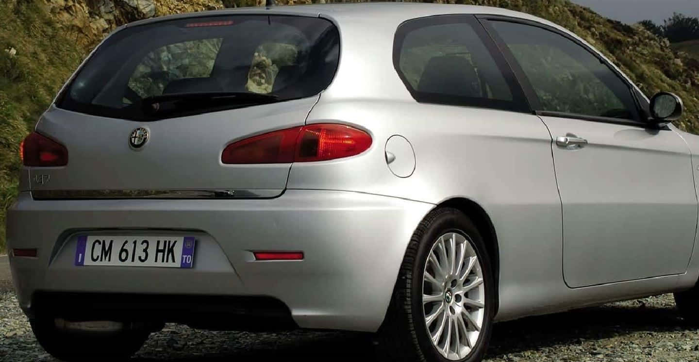 Alfa Romeo 147 桌布 壁紙 — 銀色愛快羅密歐147掀背車停在碎石上，背景是岩石山丘，天色陰沉。