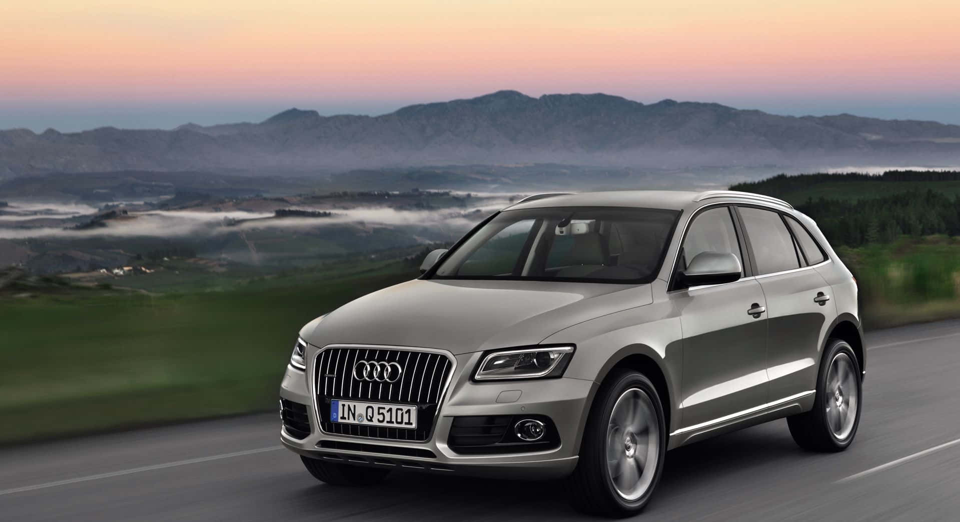 Audi Q5 桌布 壁紙 — 一輛銀色奧迪Q5 SUV在日落時分行駛在蜿蜒的道路上，背景是霧濛濛的山脈和綠色山丘。