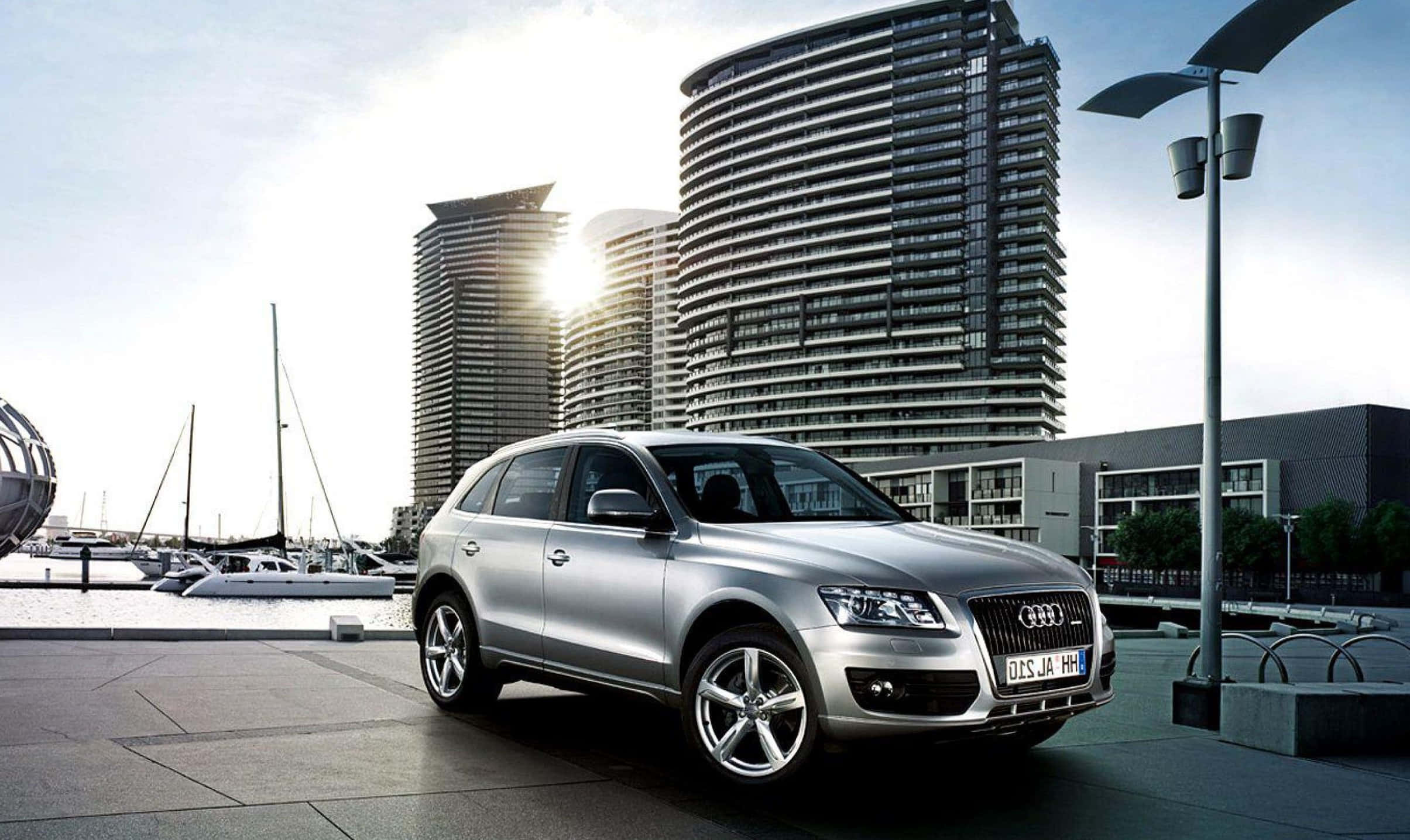 Audi Q5 桌布 壁紙 — 一輛時尚的銀色 Audi Q7 SUV 停放在現代化的海濱廣場上，背景有帆船和彎曲的摩天大樓。