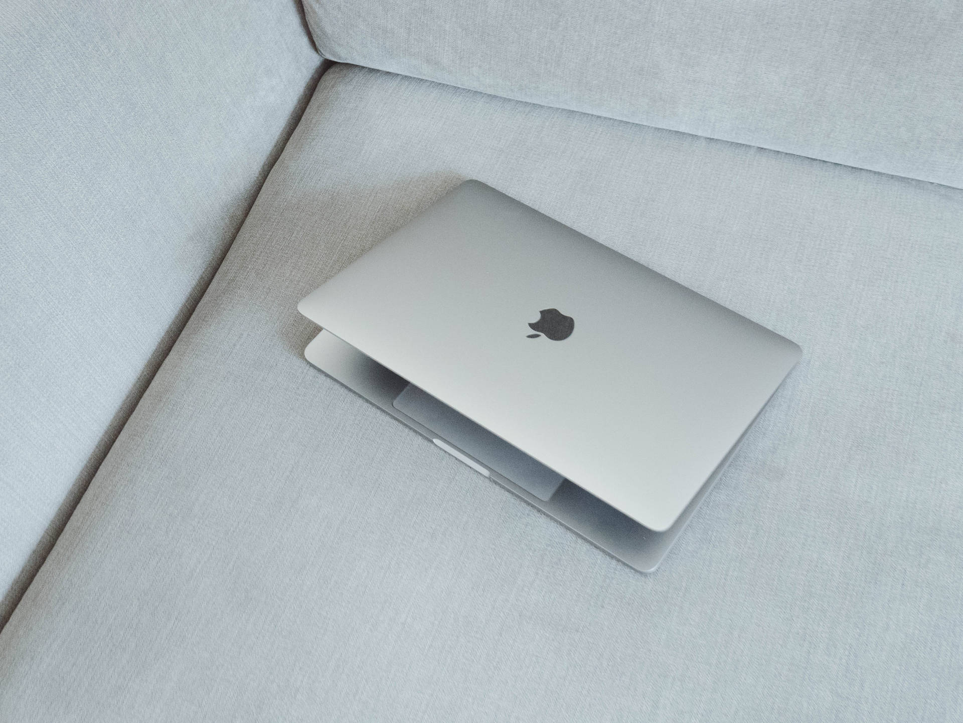 銀色筆記型電腦放在淺灰色布質沙發角落。 — Macbook Pro 壁紙