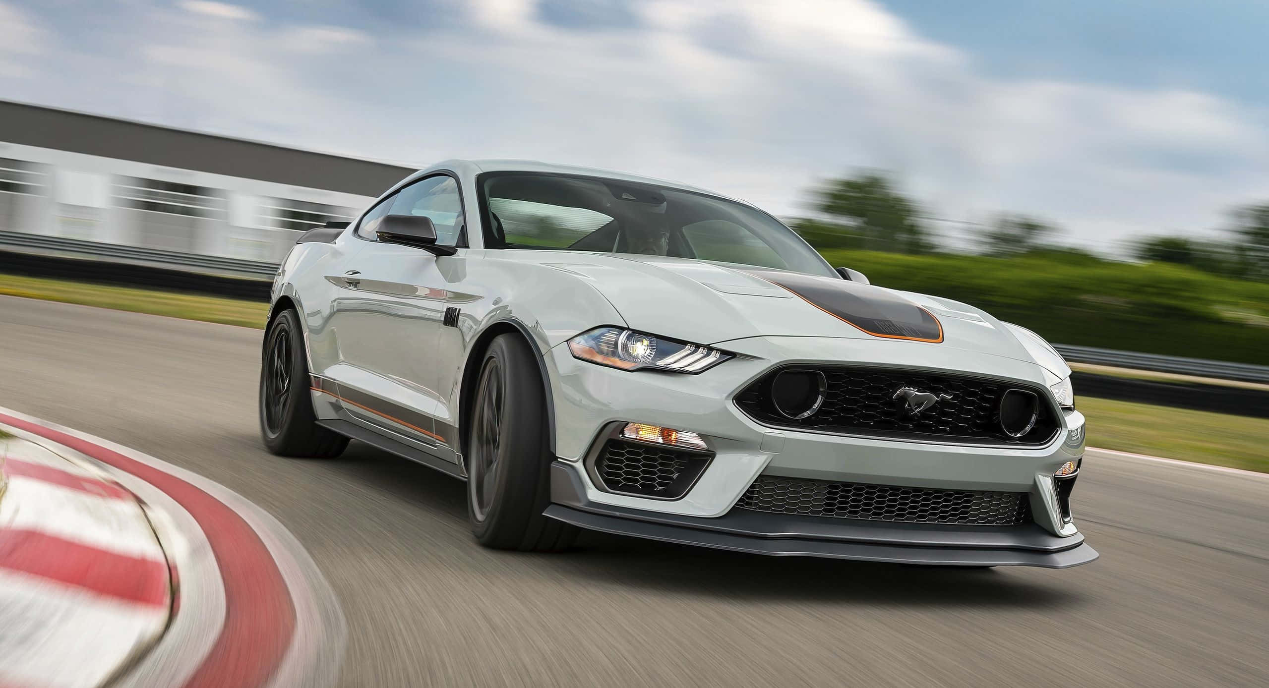 Ford Mustang Mach 1 桌布 壁紙 — 一輛銀色福特野馬，帶有黑色和橙色點綴，在部分多雲的天空下於賽道彎道高速行駛。