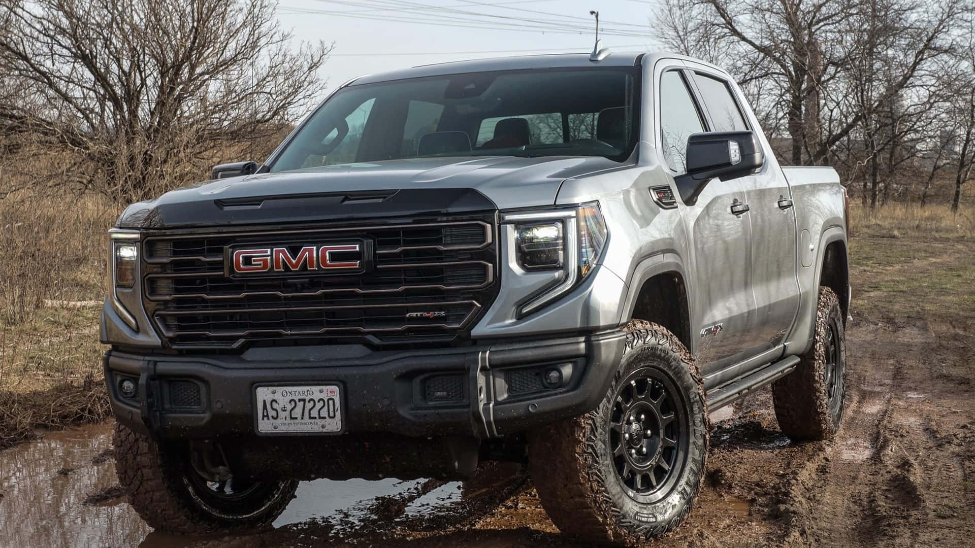 Gmc Sierra 桌布 壁紙 — 一輛銀色GMC Sierra AT4皮卡車，輪胎沾滿泥濘，駛過泥濘的田野，背景是光禿禿的樹木。