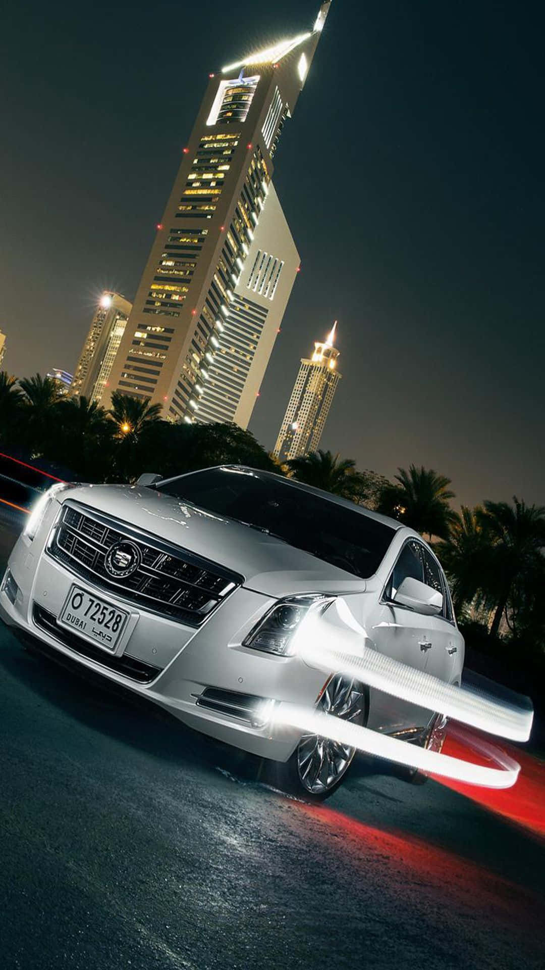 Cadillac Xts 桌布 壁紙 — 一輛銀色豪華轎車在霓虹燈閃爍的杜拜夜晚疾馳，摩天大樓和棕櫚樹在背後閃耀。