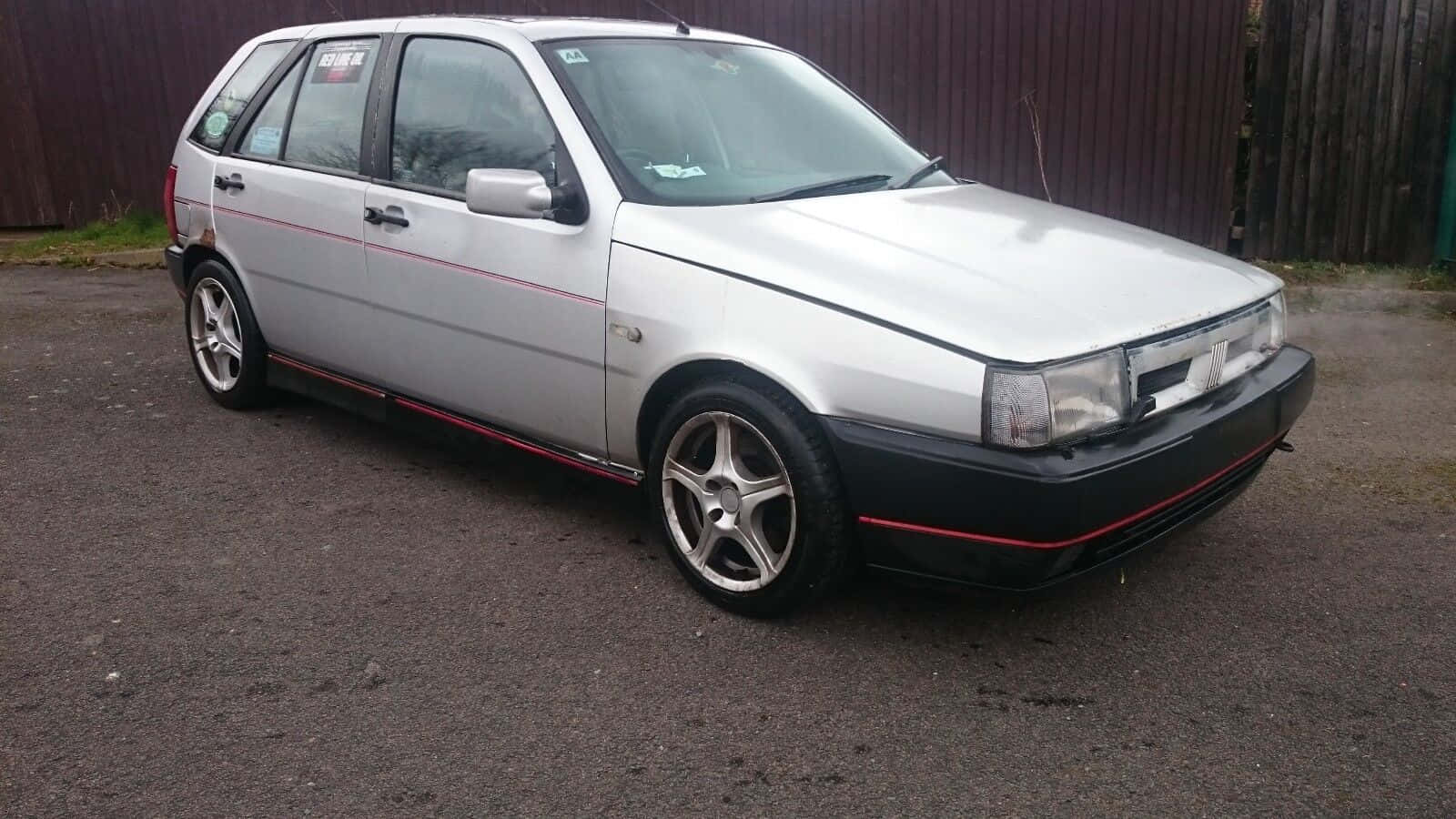 Fiat Tipo 桌布 壁紙 — 一輛銀色掀背車，帶有紅色飾條和合金輪圈，停在木柵欄附近的柏油路面上。