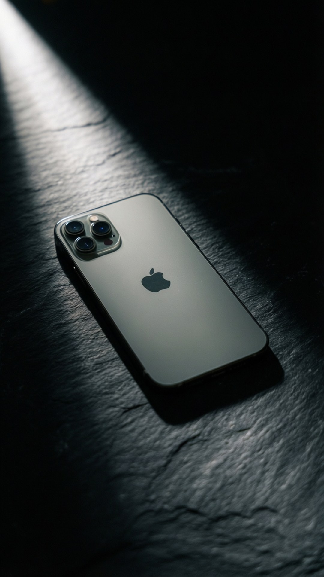 Dramatic Light iPhone 壁紙 — 一部銀色iPhone放在深色石板表面上，沐浴在單一戲劇性聚光燈下，形成深邃陰影。
