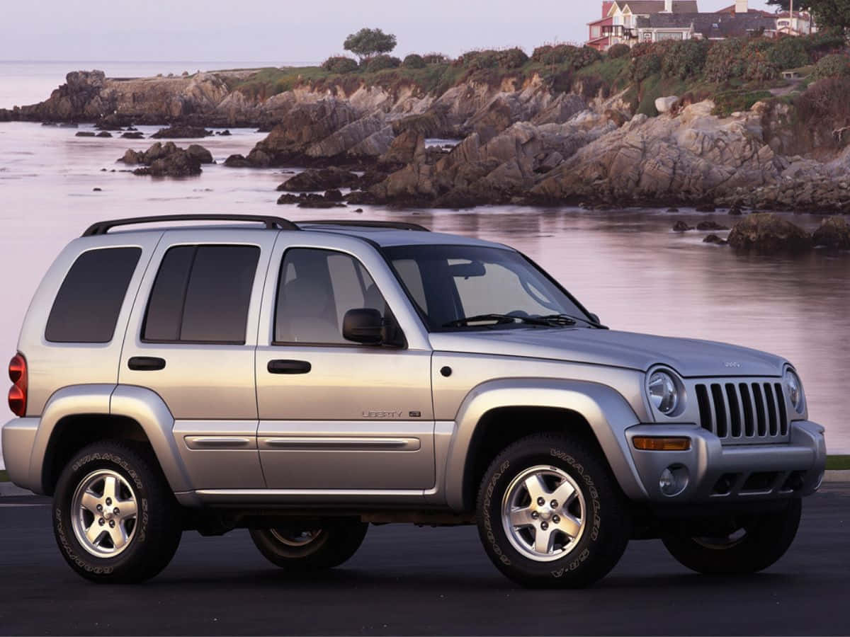 Jeep Liberty 桌布 壁紙 — 銀色Jeep Liberty SUV停泊在黃昏時分岩石海岸線旁的人行道上，懸崖上有房屋。