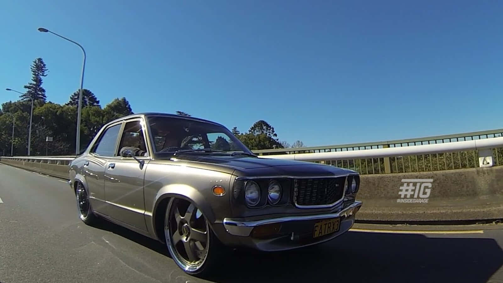 Mazda Rx-3 桌布 壁紙 — 一輛銀色經典轎車配備訂製輪圈，在晴朗藍天下行駛於陽光普照的高速公路橋上。